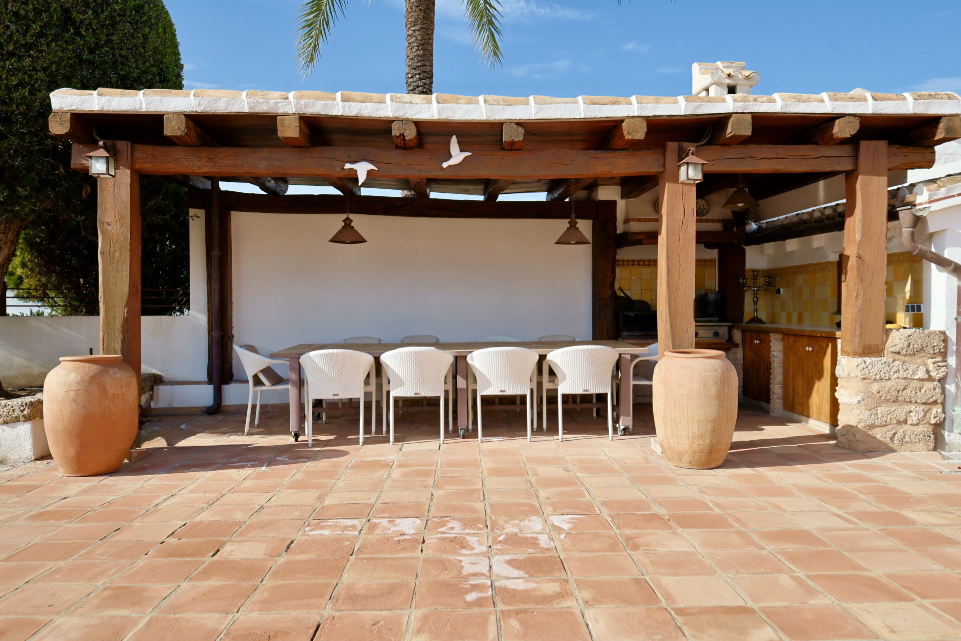 Covered outdoor dining area with long table, white wicker chairs, and terracotta tiled floor.