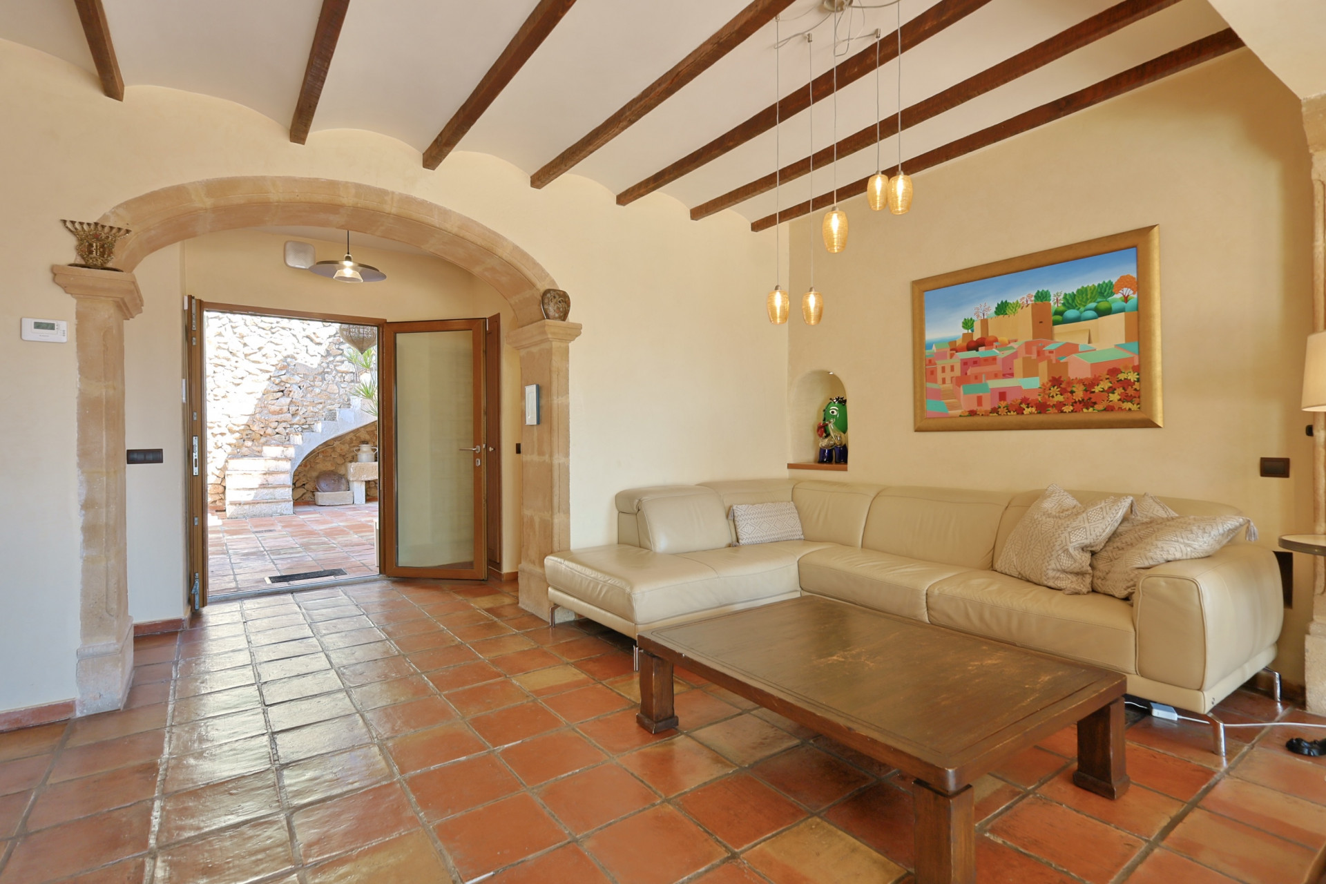 Living room with terracotta flooring, exposed wood beams, and light leather sofa. Stone archway leads to an outdoor staircase.