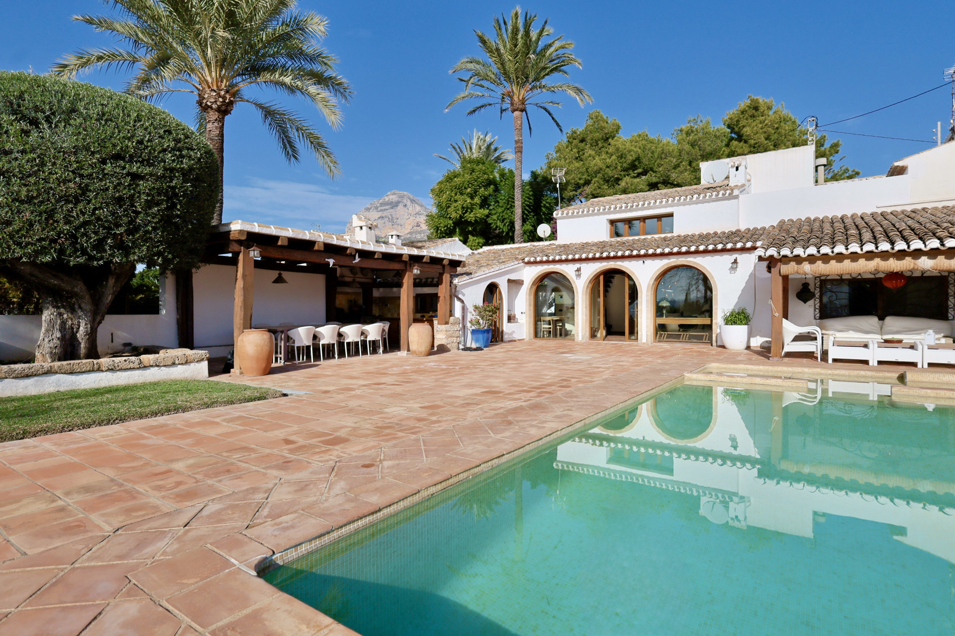 Pool view with terracotta tile, covered terrace with seating, and villa arches.