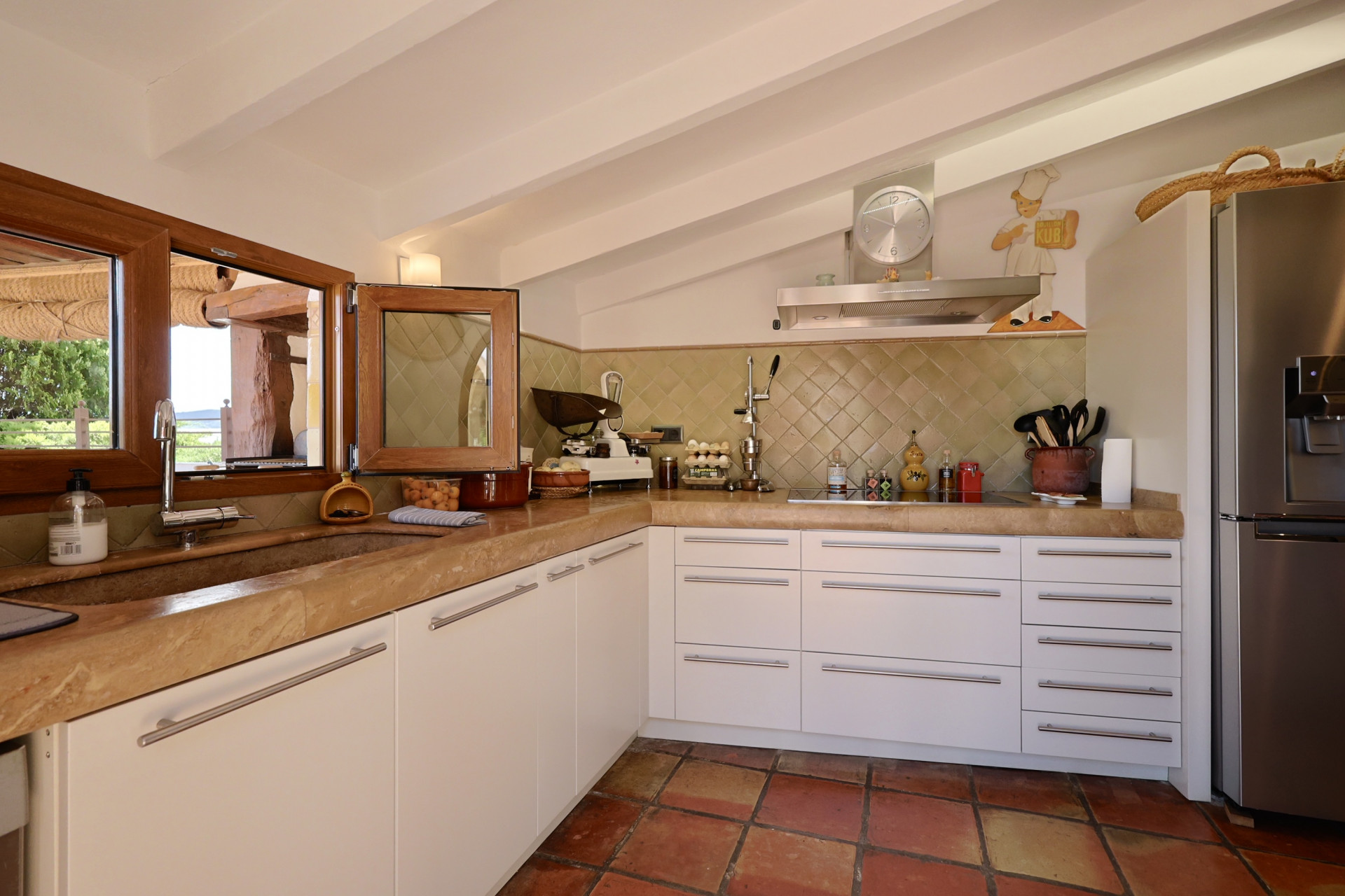 Rustic kitchen with stone countertop, white cabinets, and terracotta floor. Stone sink with wood-framed window.
