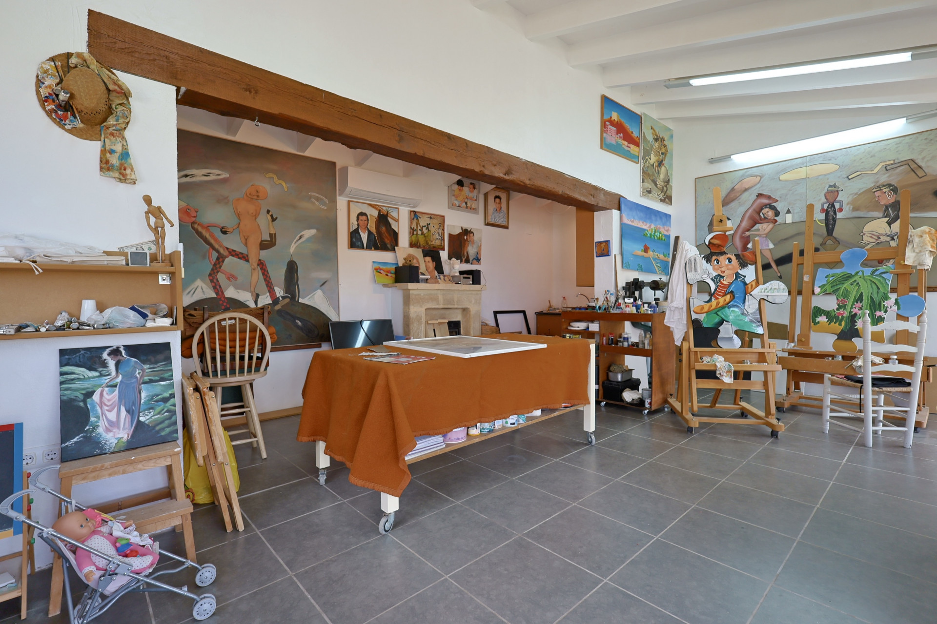 Spacious art studio featuring gray tiled floors, an orange cloth-covered table, and easels displaying paintings.