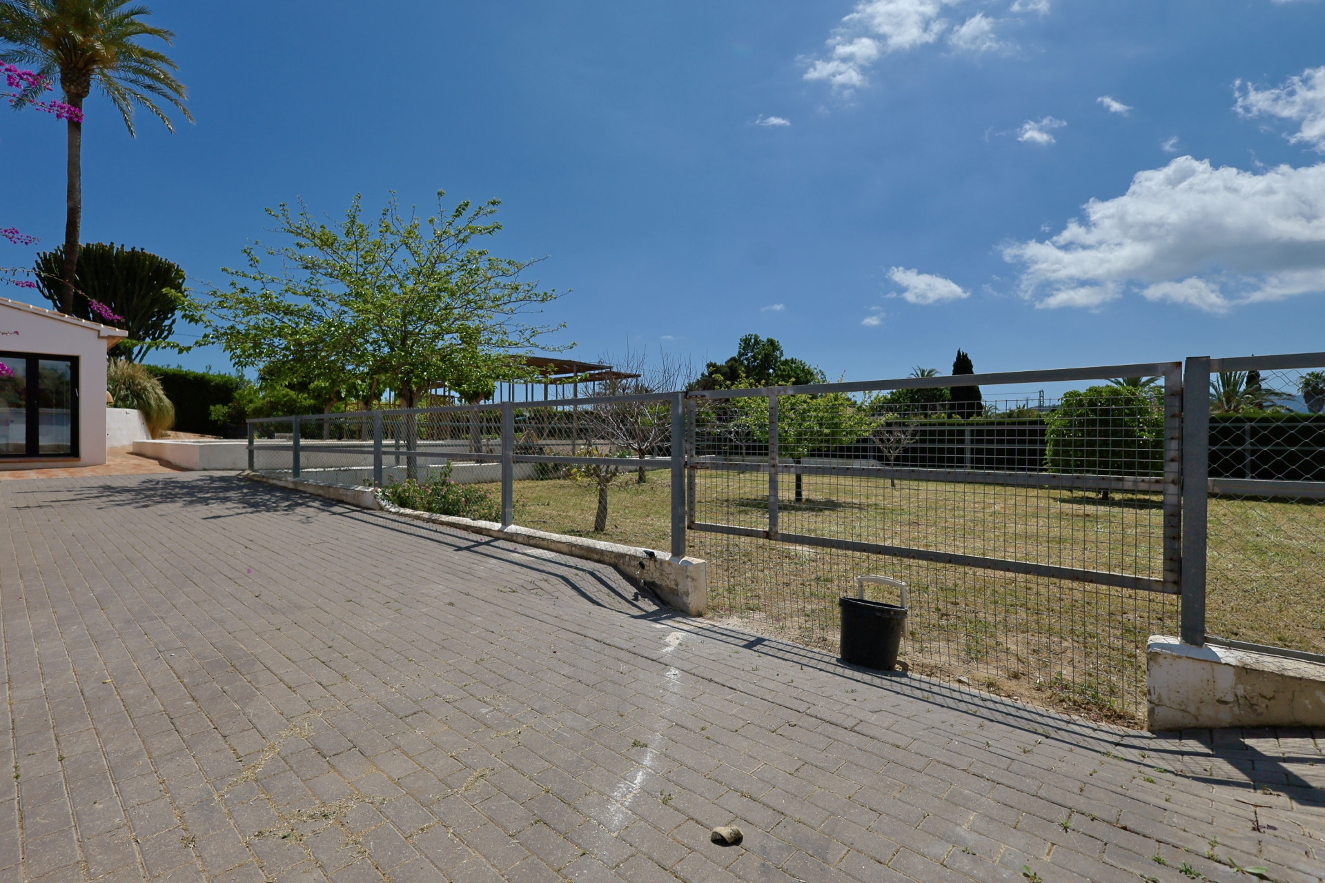 View of the fenced garden with gray pavers and lush greenery under a clear blue sky.