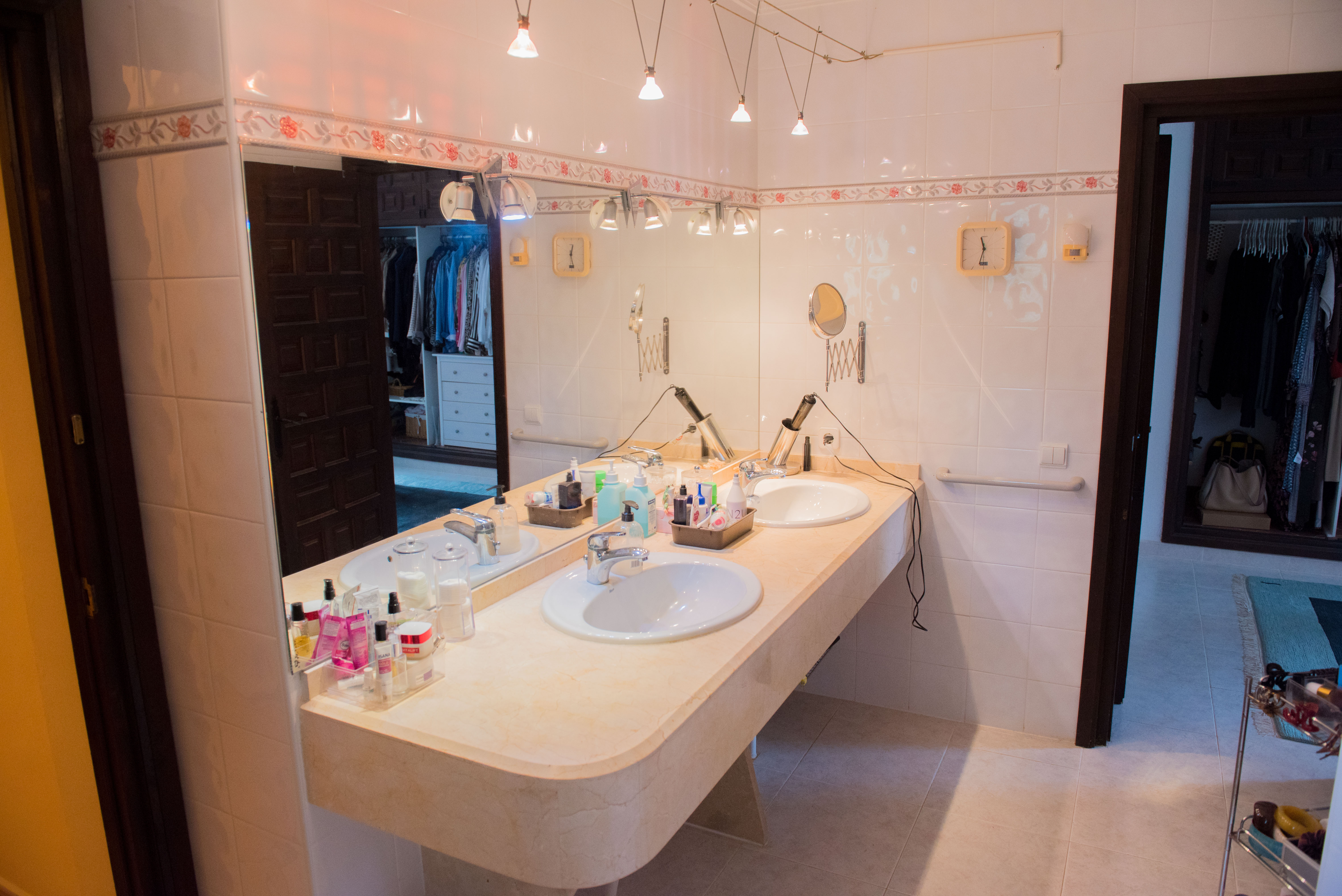 Bathroom featuring a cream marble double vanity, large mirror, and white tiled walls with floral borders, overlooking an adjacent walk-in closet.