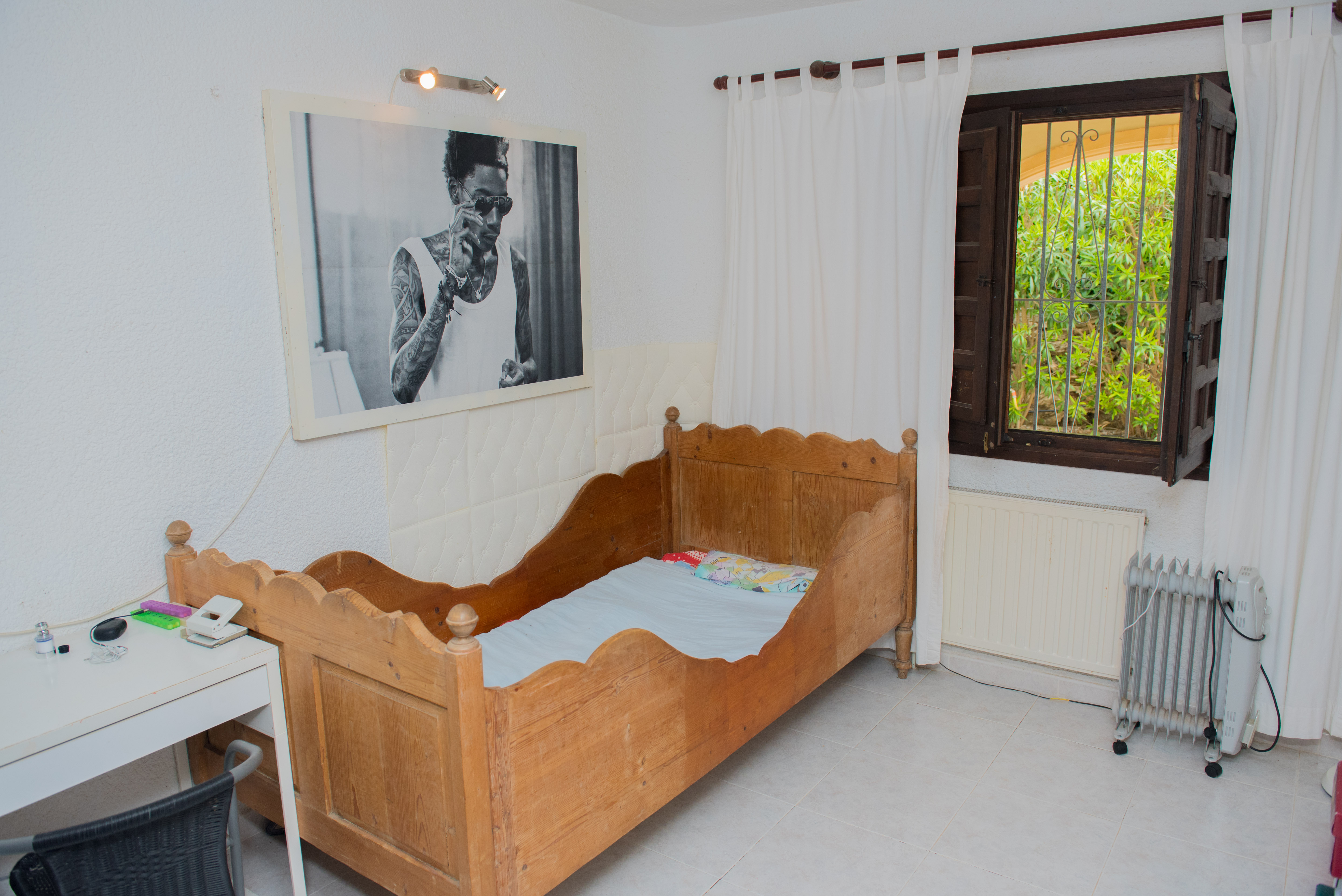 Bedroom featuring a rustic wooden bed frame with scalloped edges, a white desk, and dark wood window. The space includes tiled flooring and garden views.