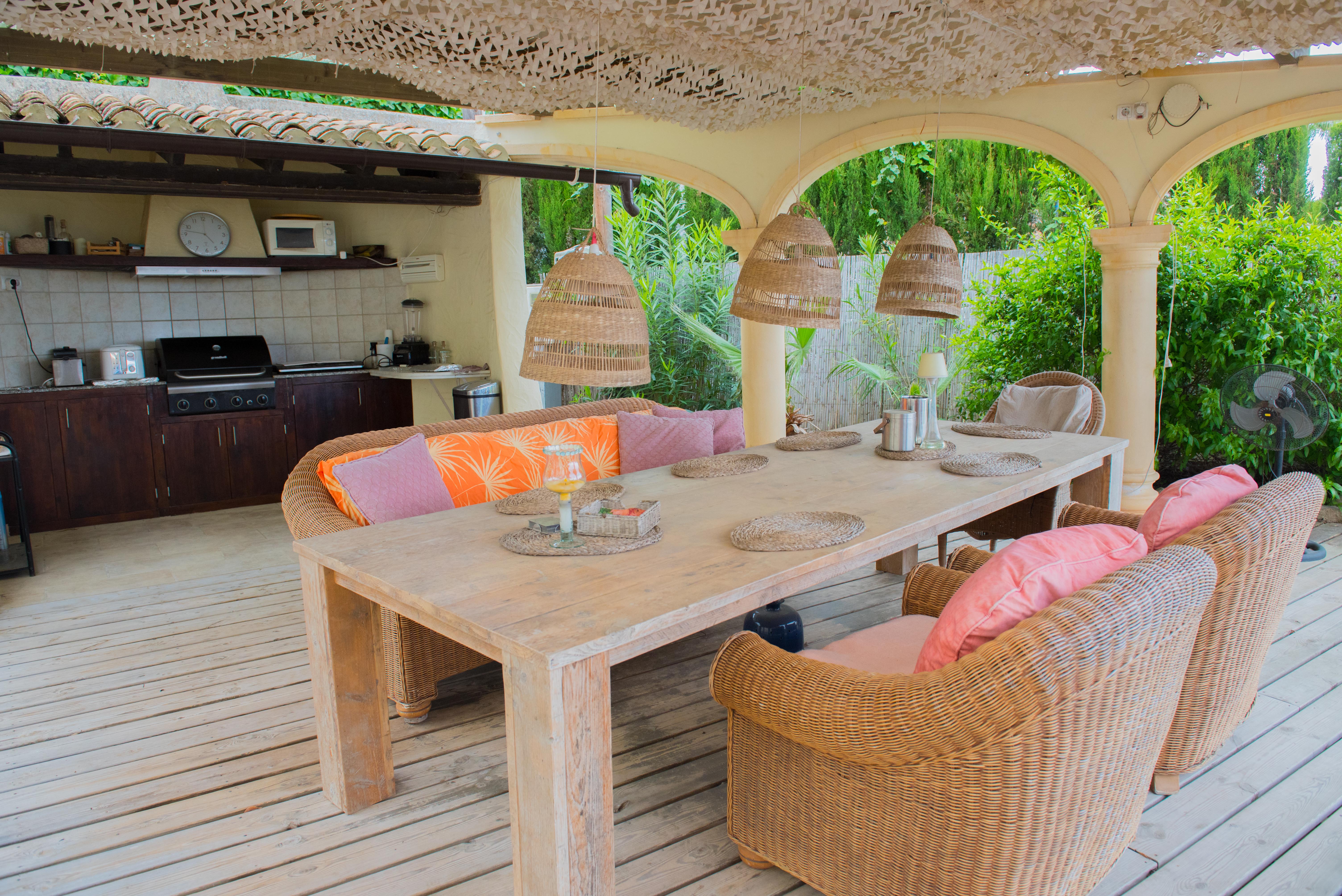 Covered terrace featuring a fully equipped summer kitchen, solid wood dining table, wicker armchairs, and decorative shading over wooden decking.