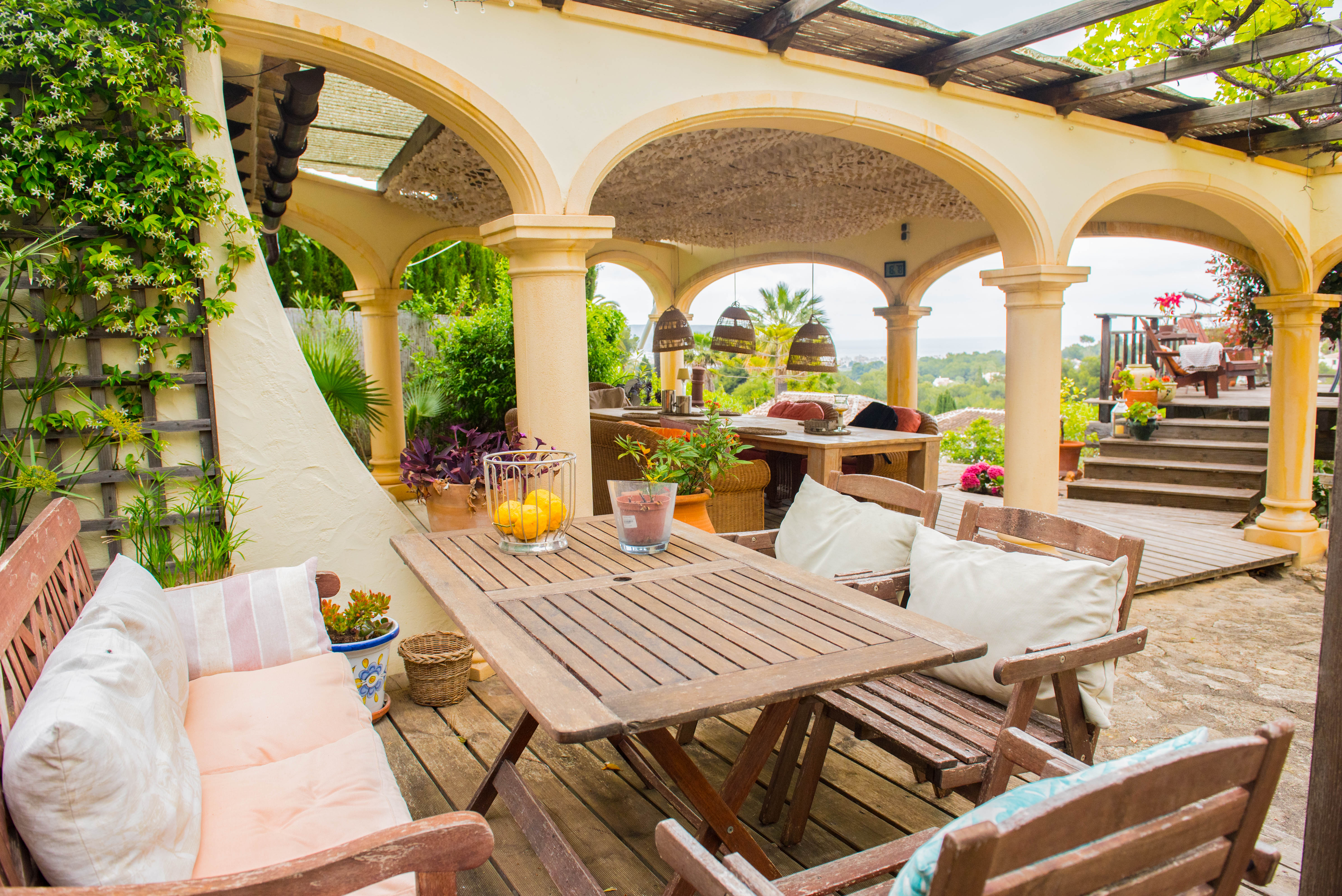 Covered terrace featuring Mediterranean arches, wooden dining furniture, timber decking, and panoramic views of the sea and lush landscape.