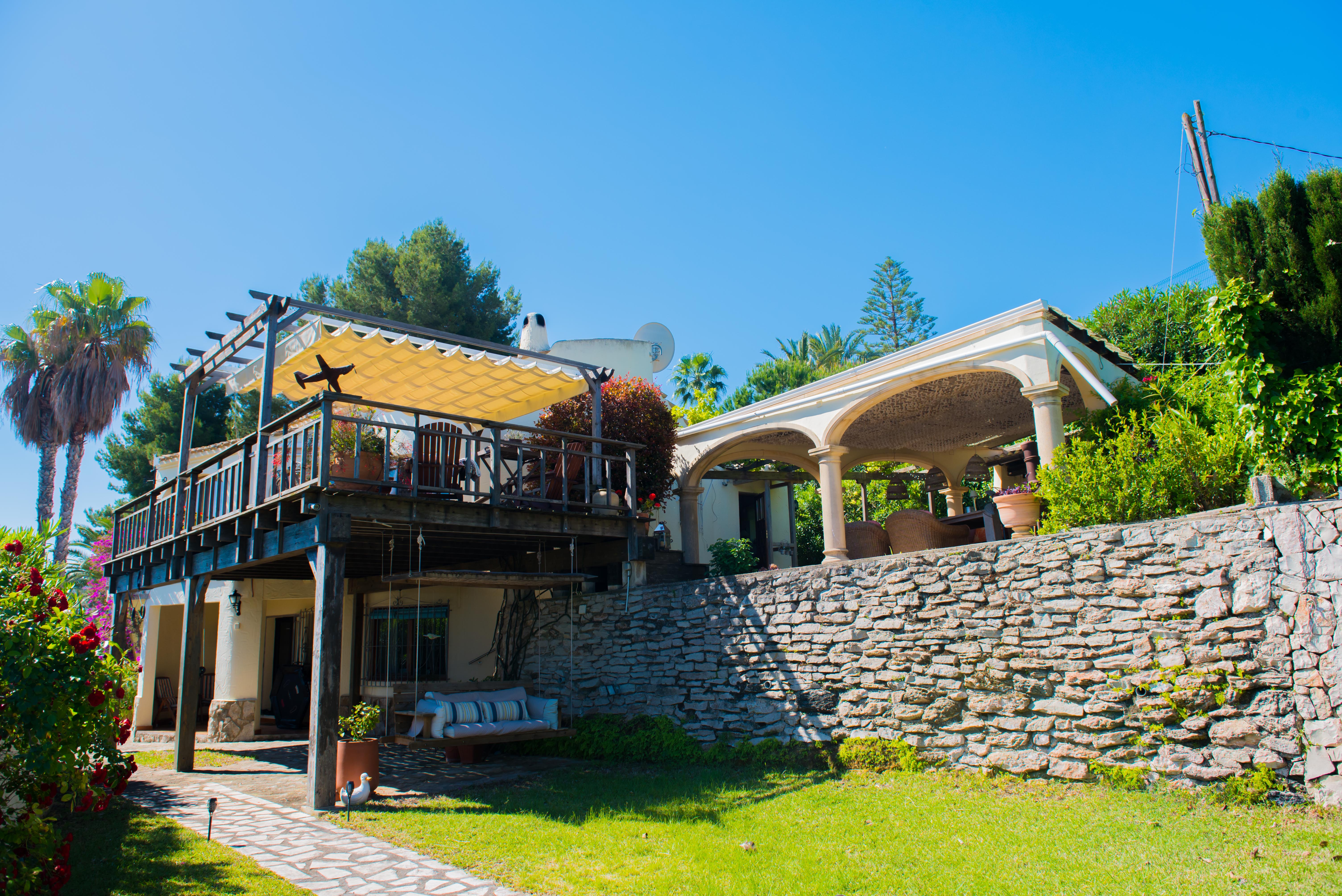 Exterior view of the villa with a wooden covered terrace and awning, garden with stone wall, and relaxation area. Exterior view of the villa with a wooden covered terrace and awning, garden with stone wall, and relaxation area.