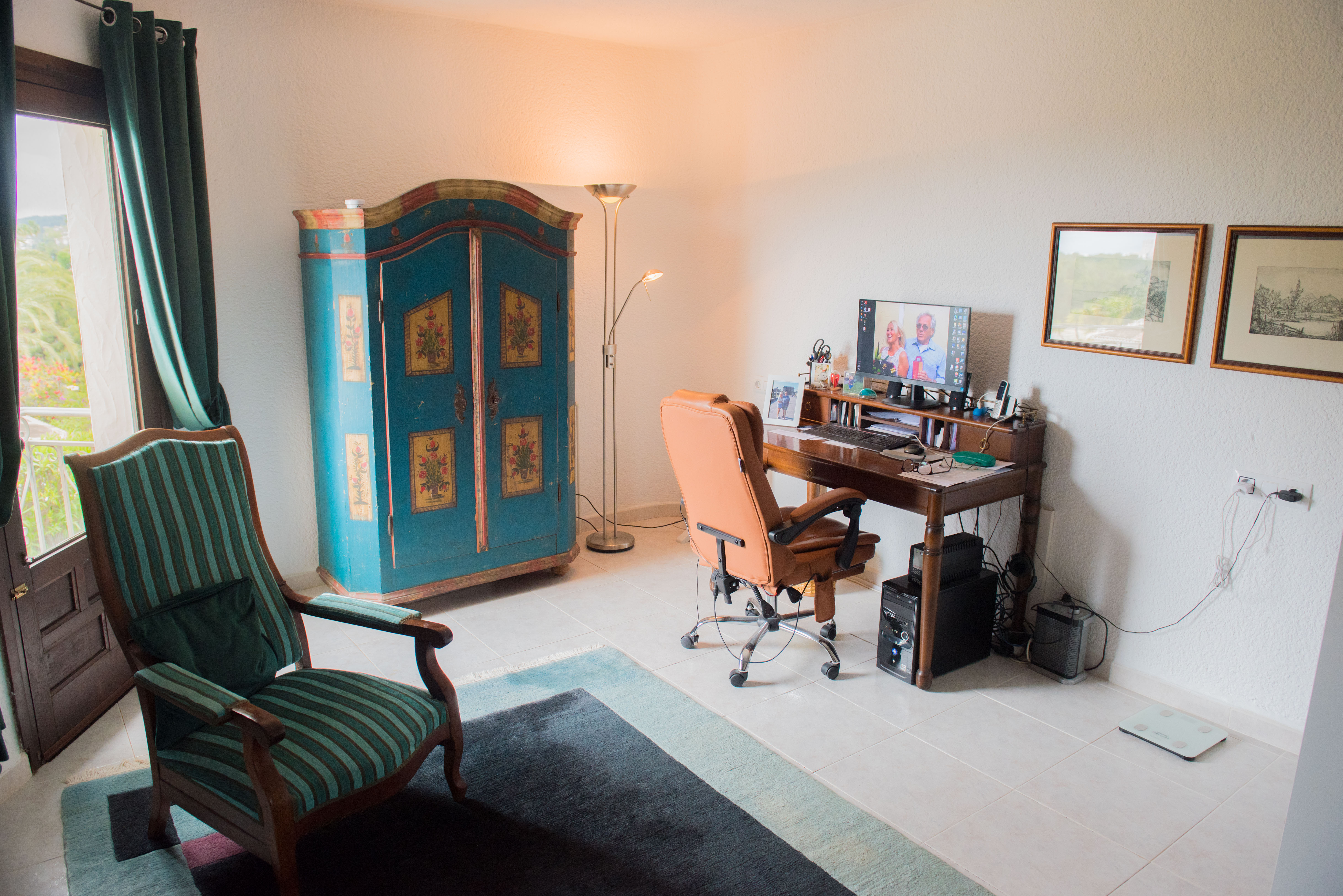 Functional home office with a classic wooden desk, decorated rustic armoire, and leather chair. Features natural light and direct terrace access.