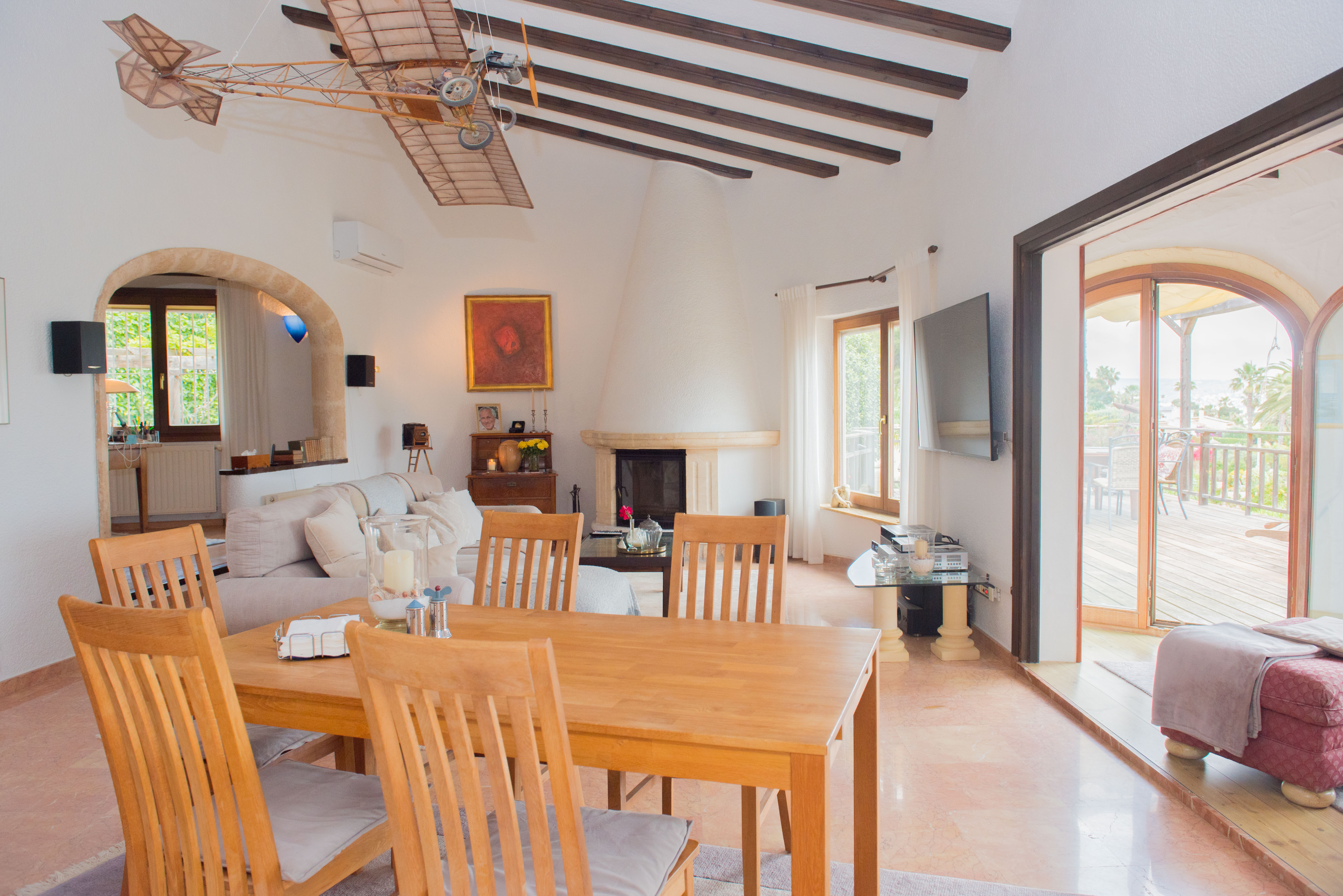 Living and dining area featuring exposed wooden beams, a conical white fireplace, and direct access to a wooden terrace through large arched doors.