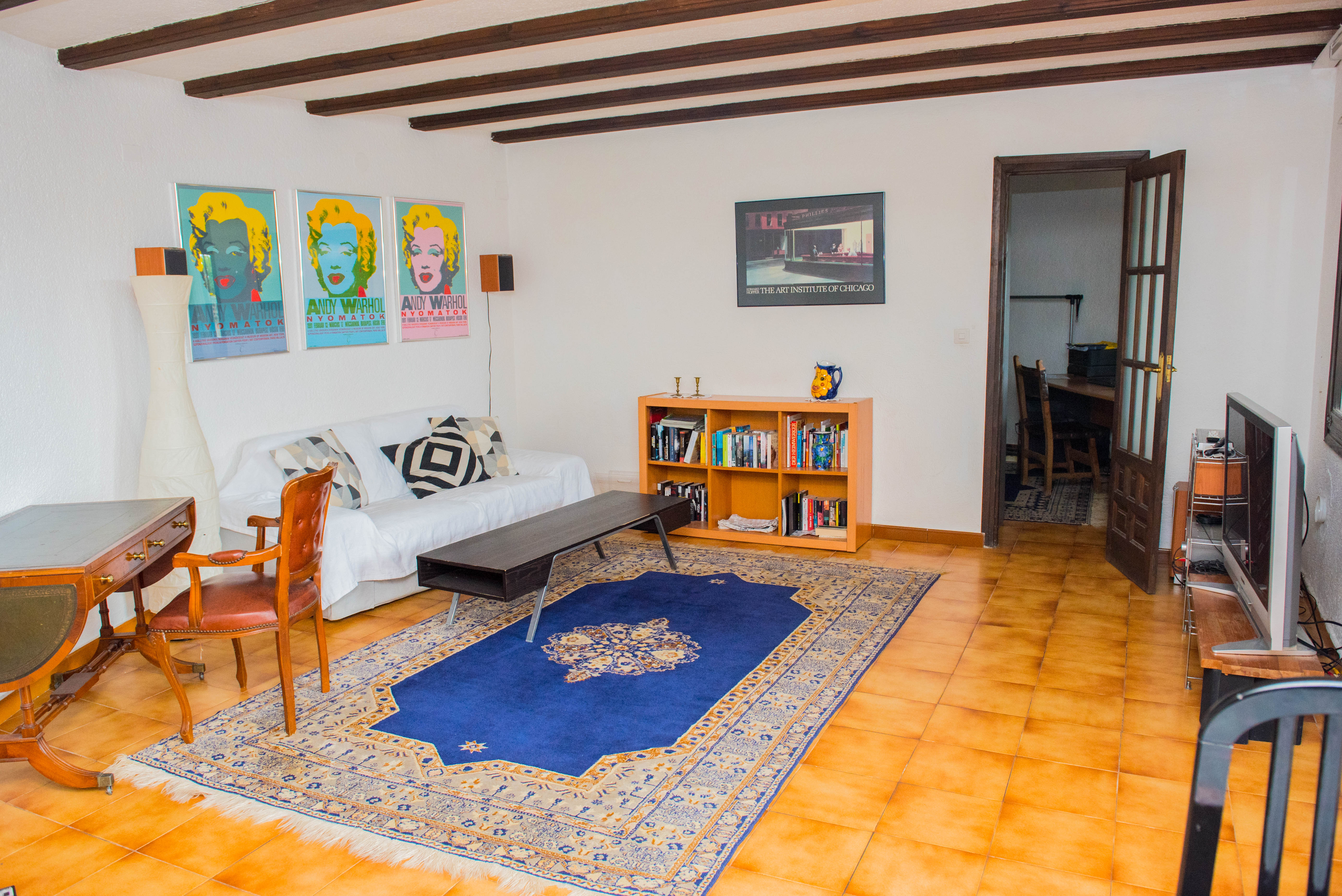 Living area featuring exposed dark wood ceiling beams, terracotta tile flooring, a white sofa, classic wooden desk, and a blue patterned rug.