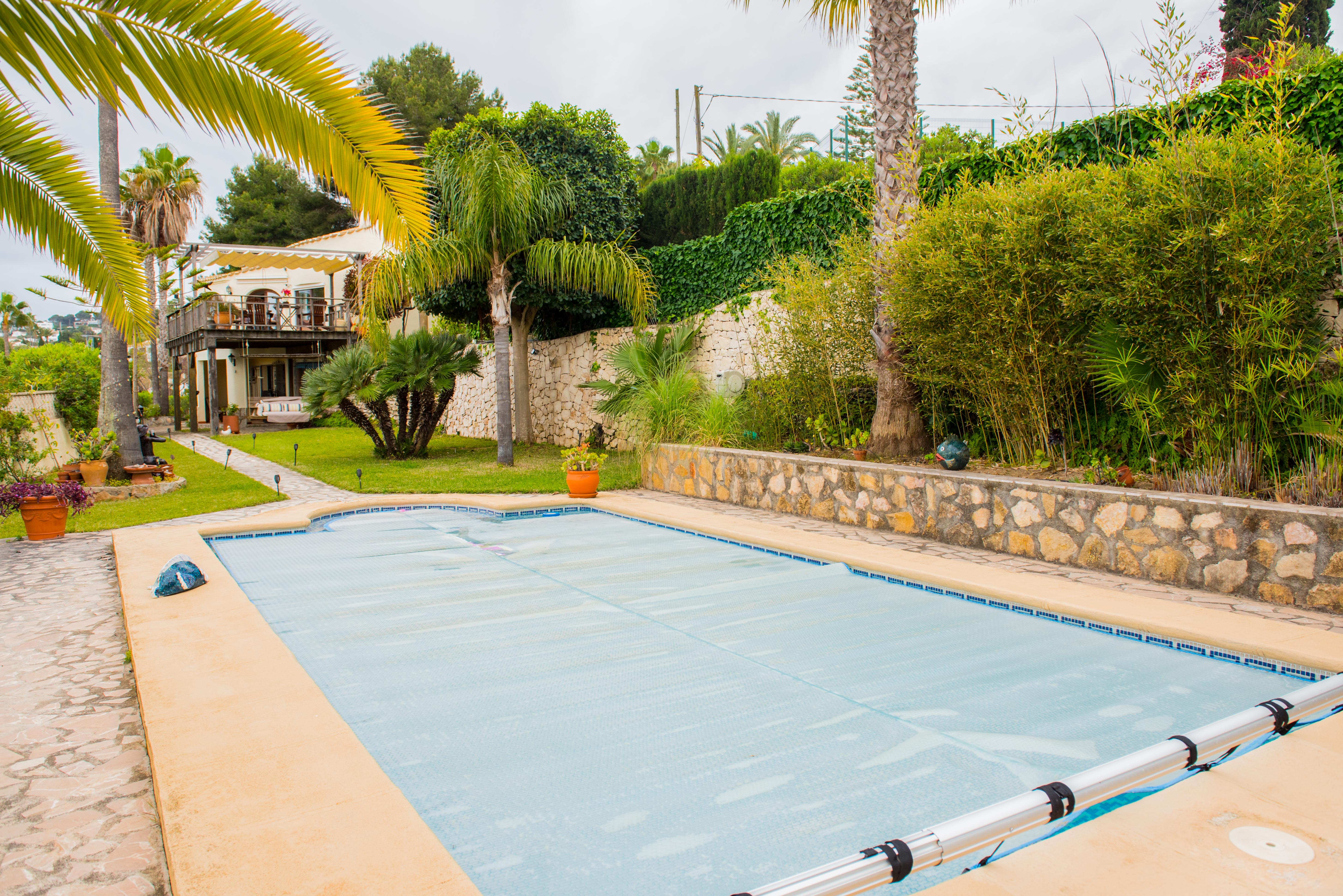 Outdoor swimming pool featuring a blue thermal cover and manual roller, set within a landscaped garden with natural stone walls, mature palms, and lush bamboo.