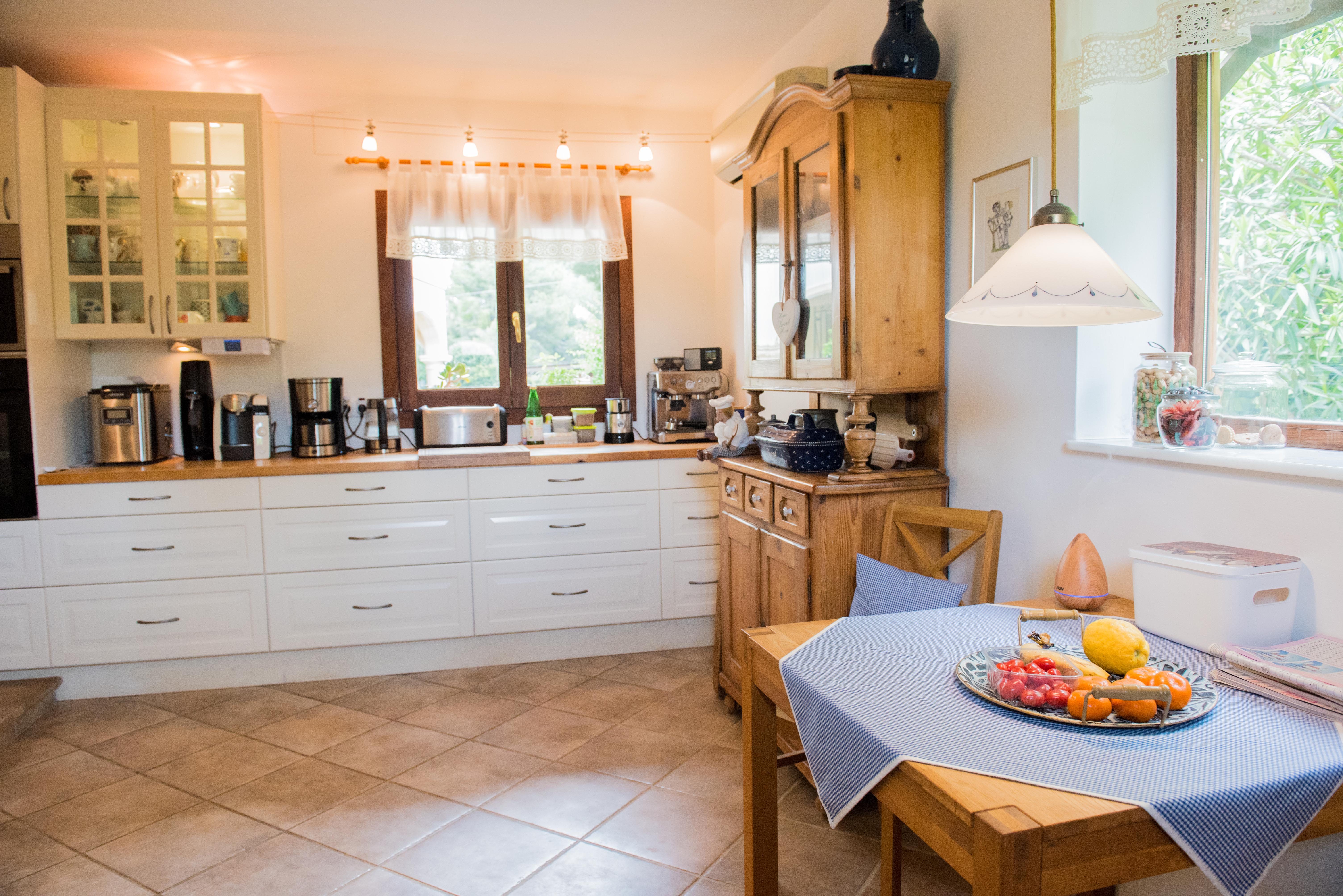 Rustic kitchen featuring white cabinetry, wooden countertops, and a dining area. It includes a classic wooden hutch, ceramic tile flooring, and natural light from two windows.