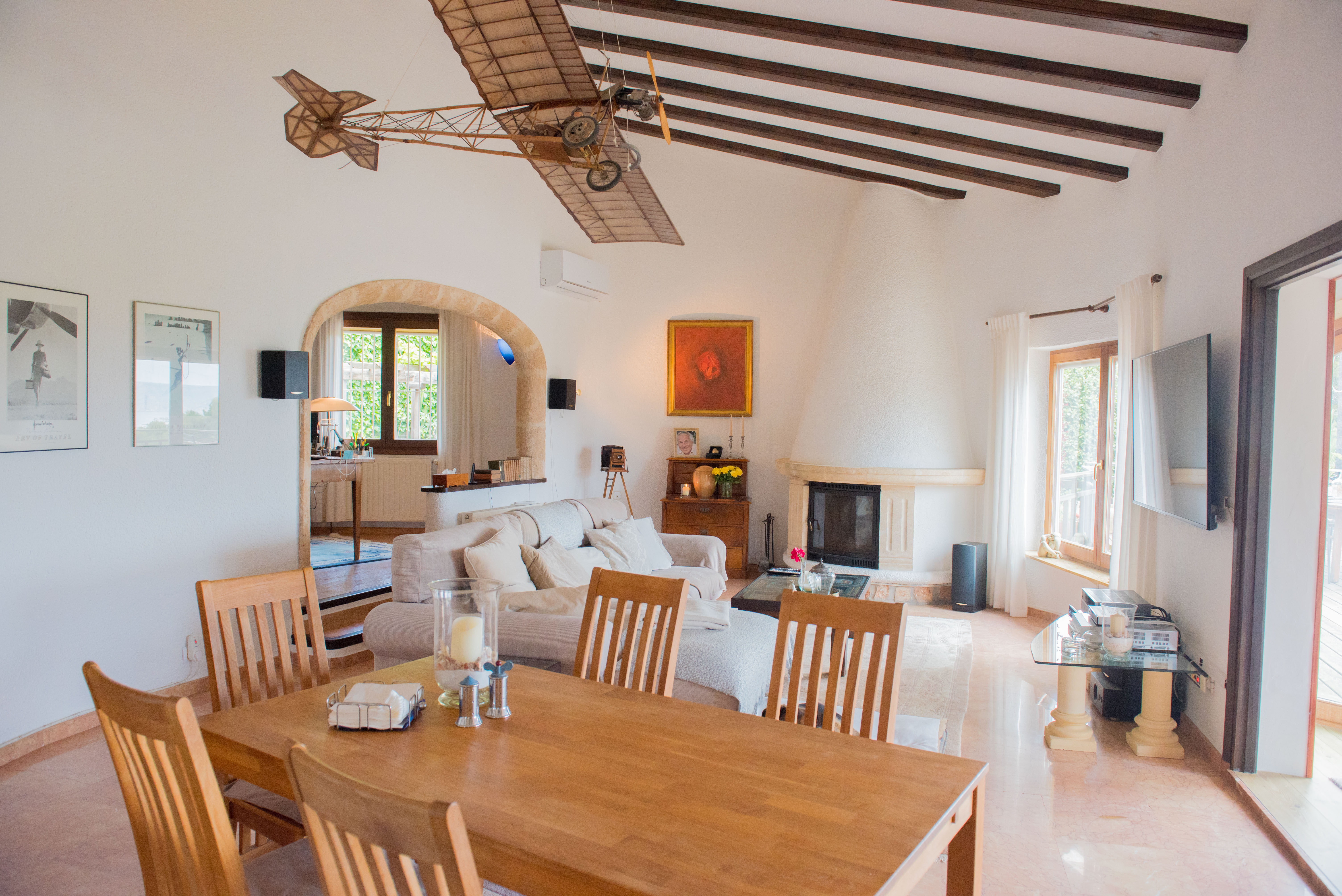 Spacious living-dining room with exposed dark wood beams, a conical fireplace, and a stone archway. Features include marble flooring and ample natural light from the terrace.