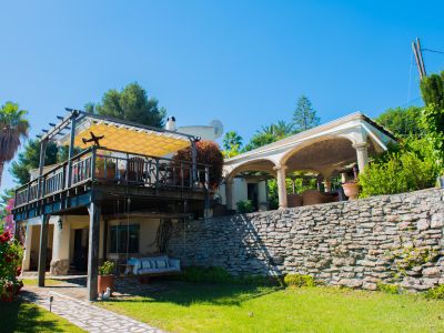 Exterior view of the villa with a wooden covered terrace and awning, garden with stone wall, and relaxation area.