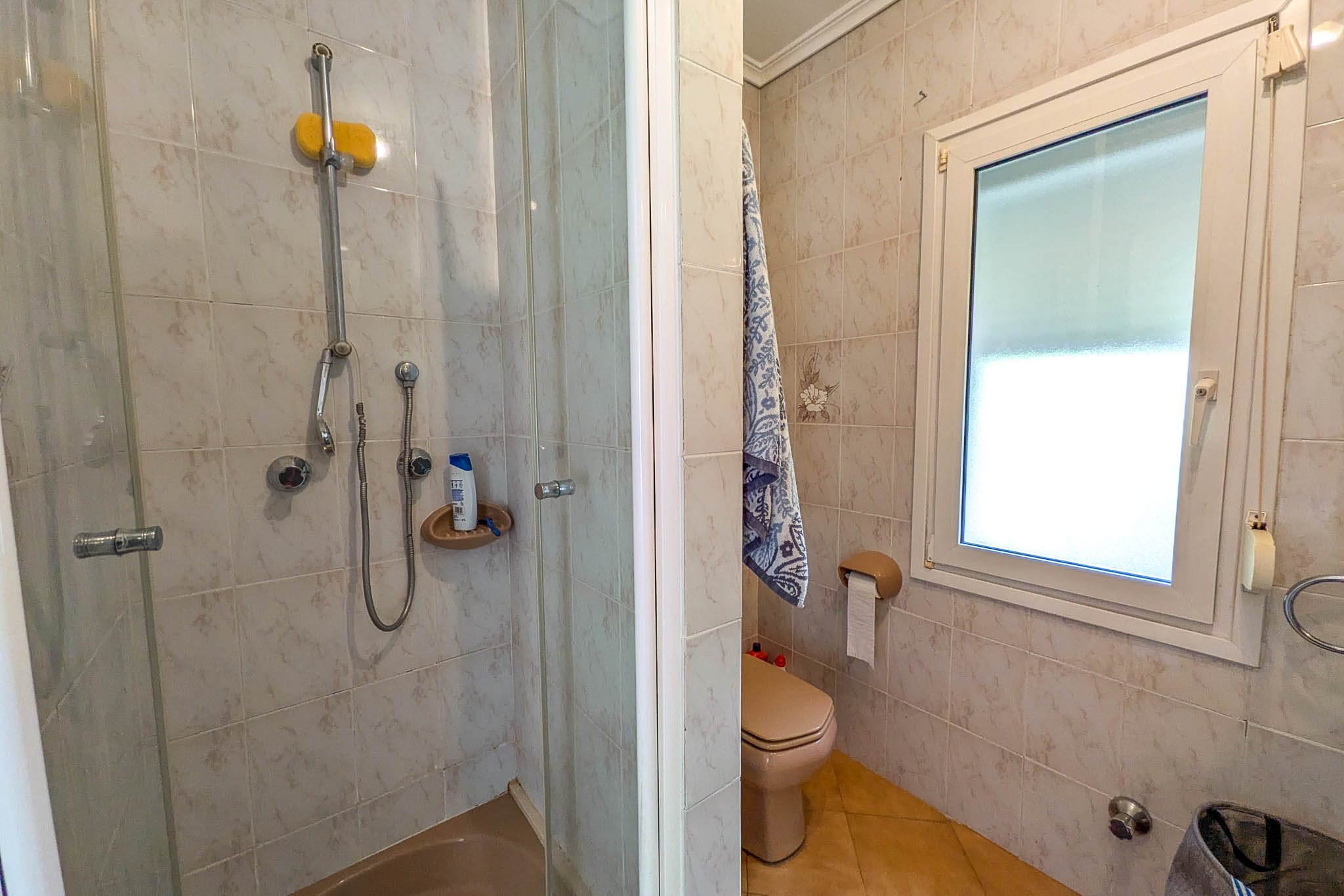 Bathroom featuring a walk-in glass shower, cream-colored toilet, and a white PVC window for natural ventilation. The walls are finished with patterned ceramic tiles.