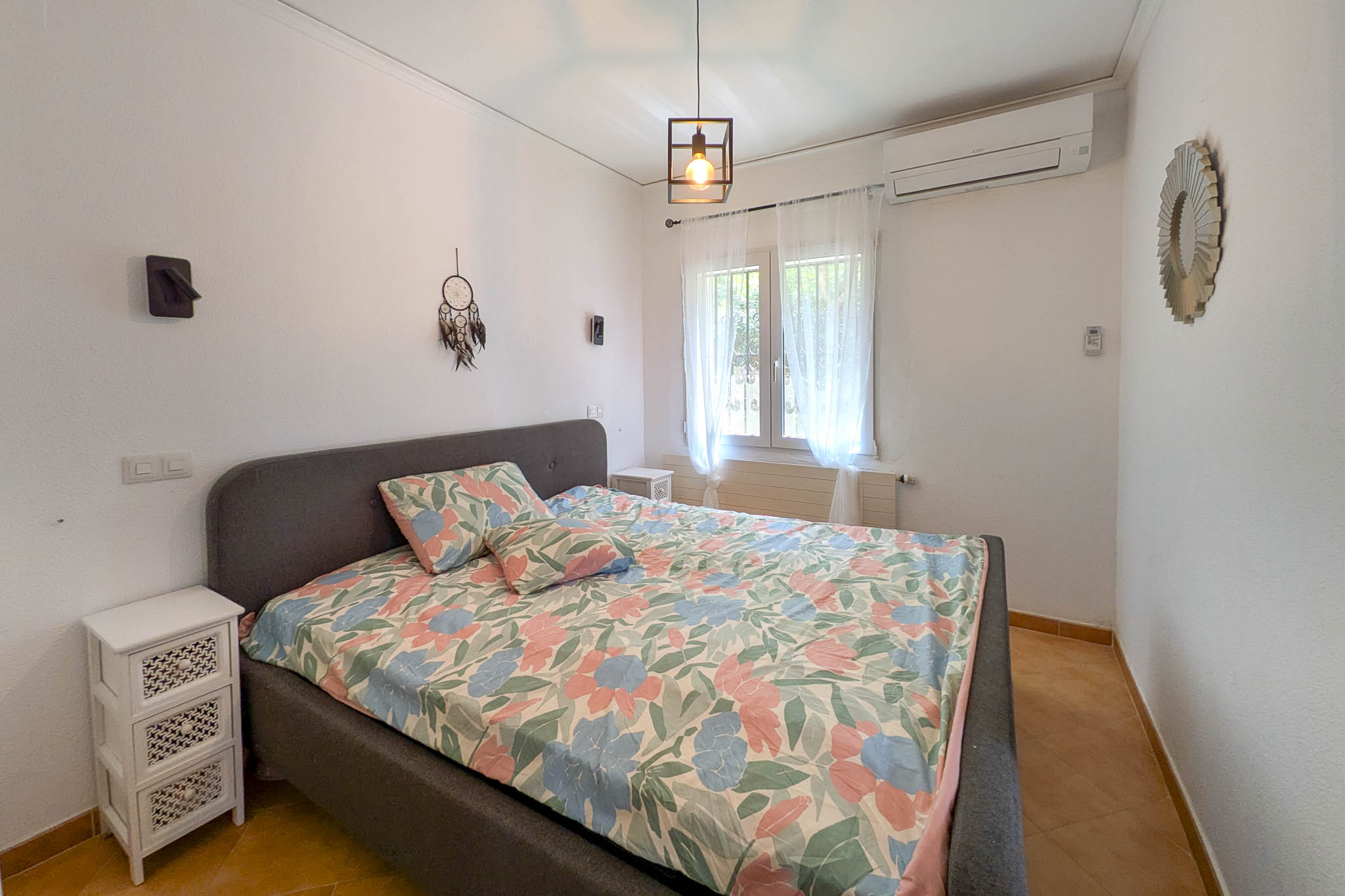 Bedroom featuring a grey upholstered bed frame, split air conditioning, and terracotta tile flooring. The space includes a window with natural light and industrial-style pendant lighting.