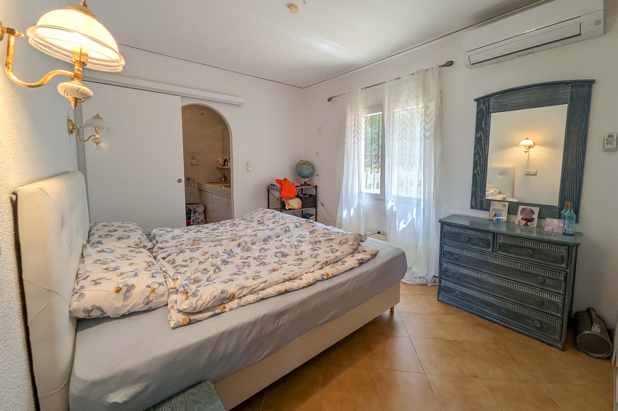 Bedroom featuring an en-suite bathroom, split air conditioning, terracotta tile flooring, and wooden furniture. The space offers natural light through lace curtains.