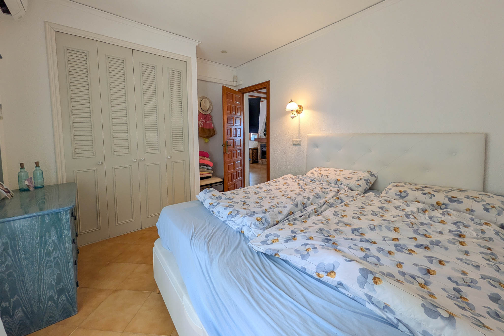 Bedroom featuring built-in wardrobes with louvred doors, terracotta-style flooring, and a tufted white headboard. A blue wooden dresser complements the neutral walls.