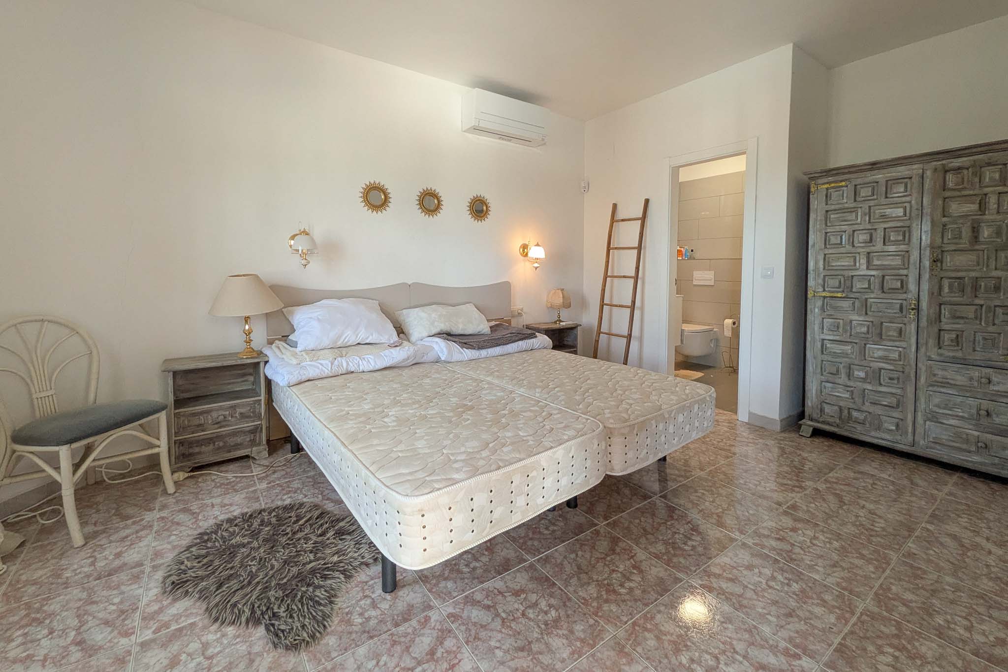 Bedroom with en-suite bathroom, marble-effect tile flooring, rustic carved wooden wardrobe, and wall-mounted air conditioning unit.