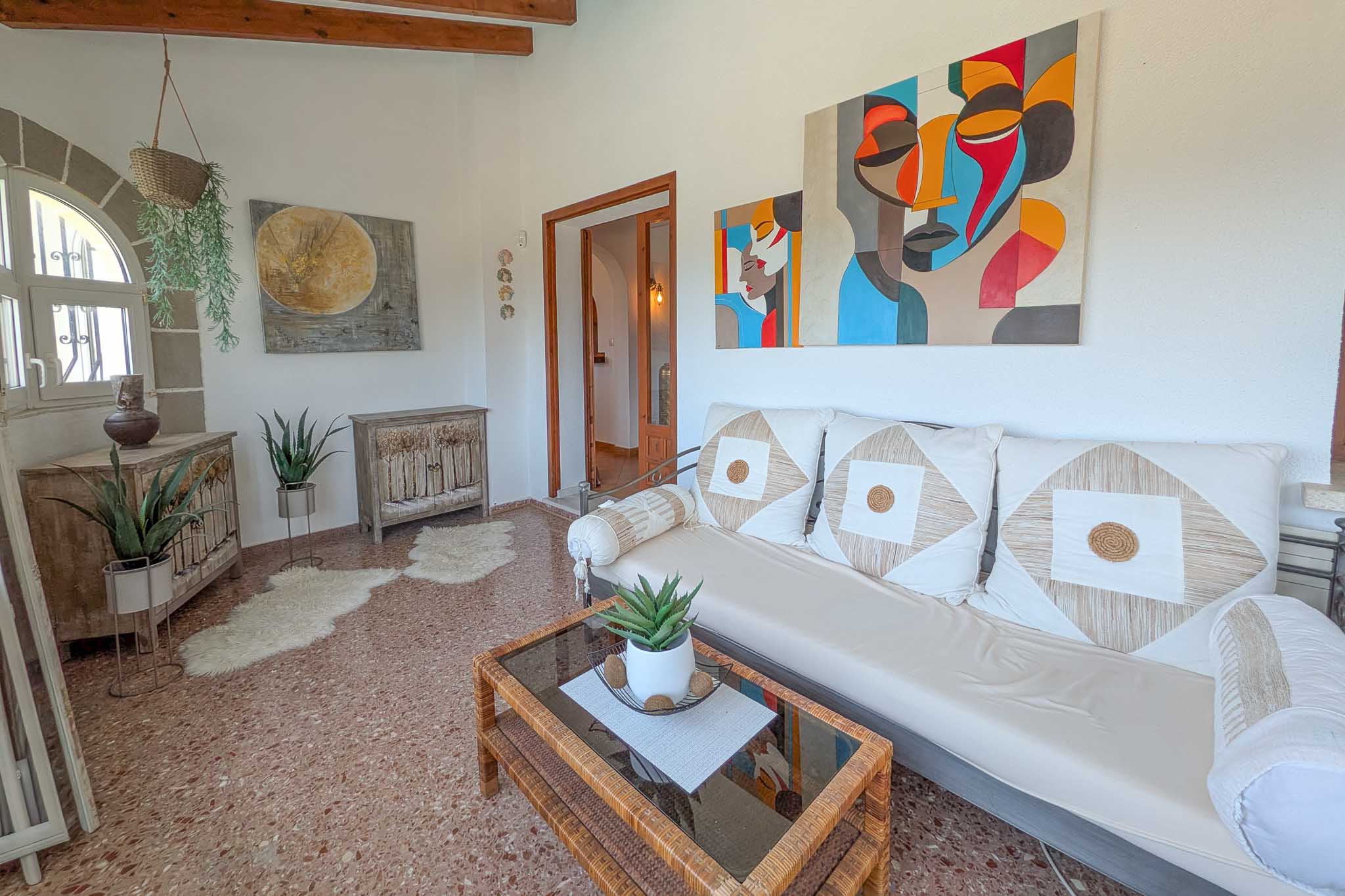 Bright living area featuring exposed wooden beams, terrazzo flooring, and an arched window. The space blends rustic wooden furniture with modern art and natural light.