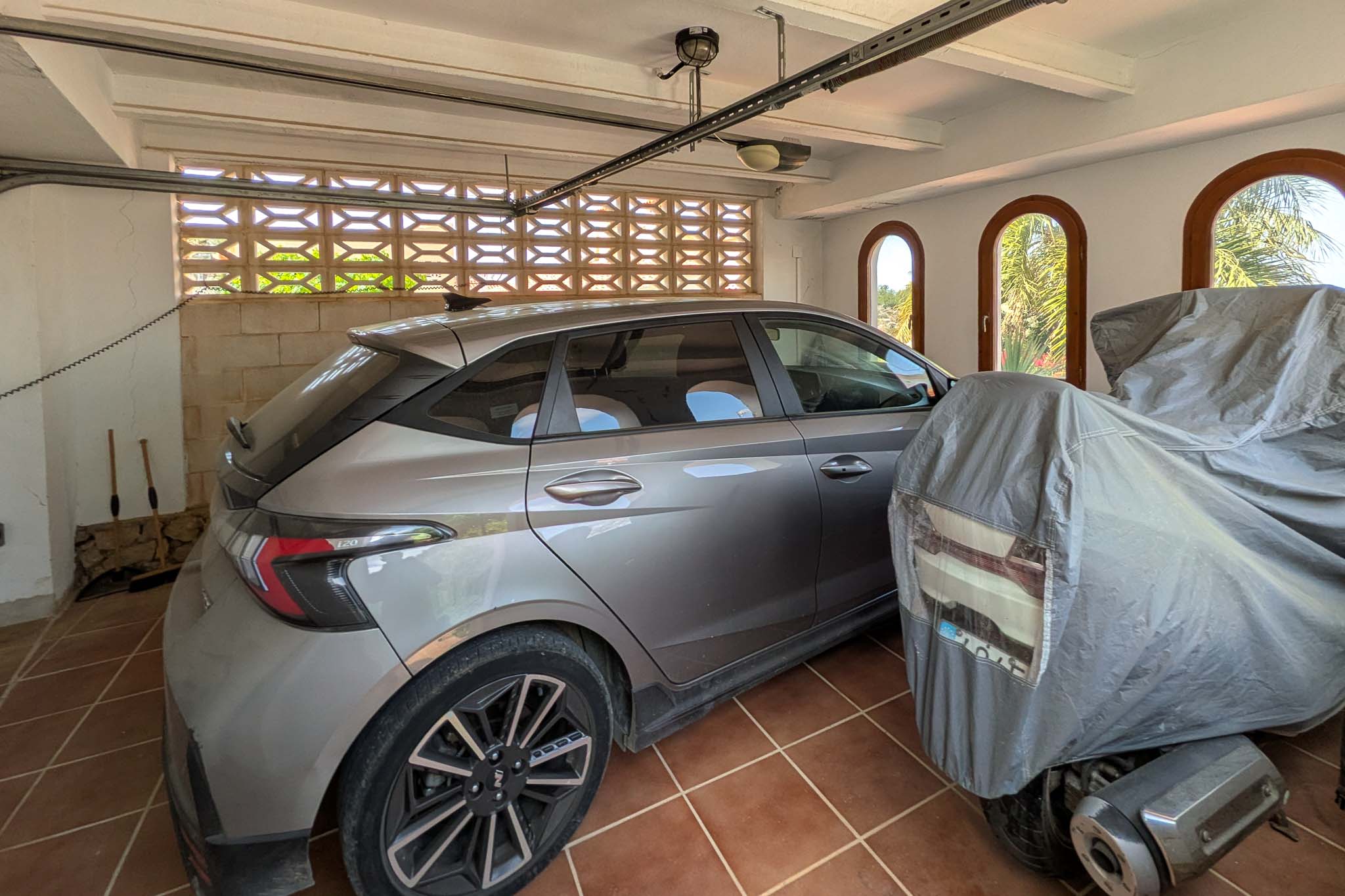 Covered garage featuring terracotta tiled flooring, white beamed ceilings, and arched windows. A decorative lattice wall provides natural ventilation for the parking area.