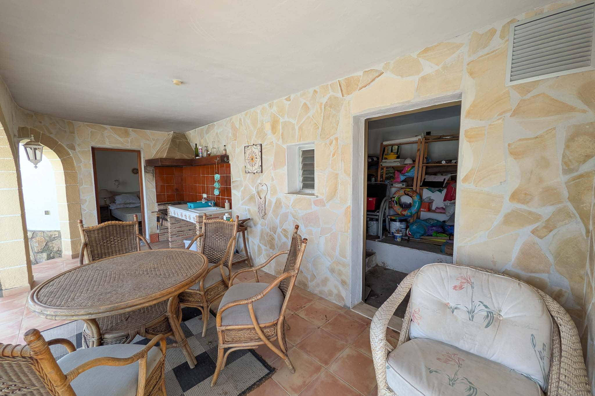 Covered ground-floor terrace featuring natural stone walls, a built-in summer kitchen, and a wicker dining area adjacent to the versatile storage room.