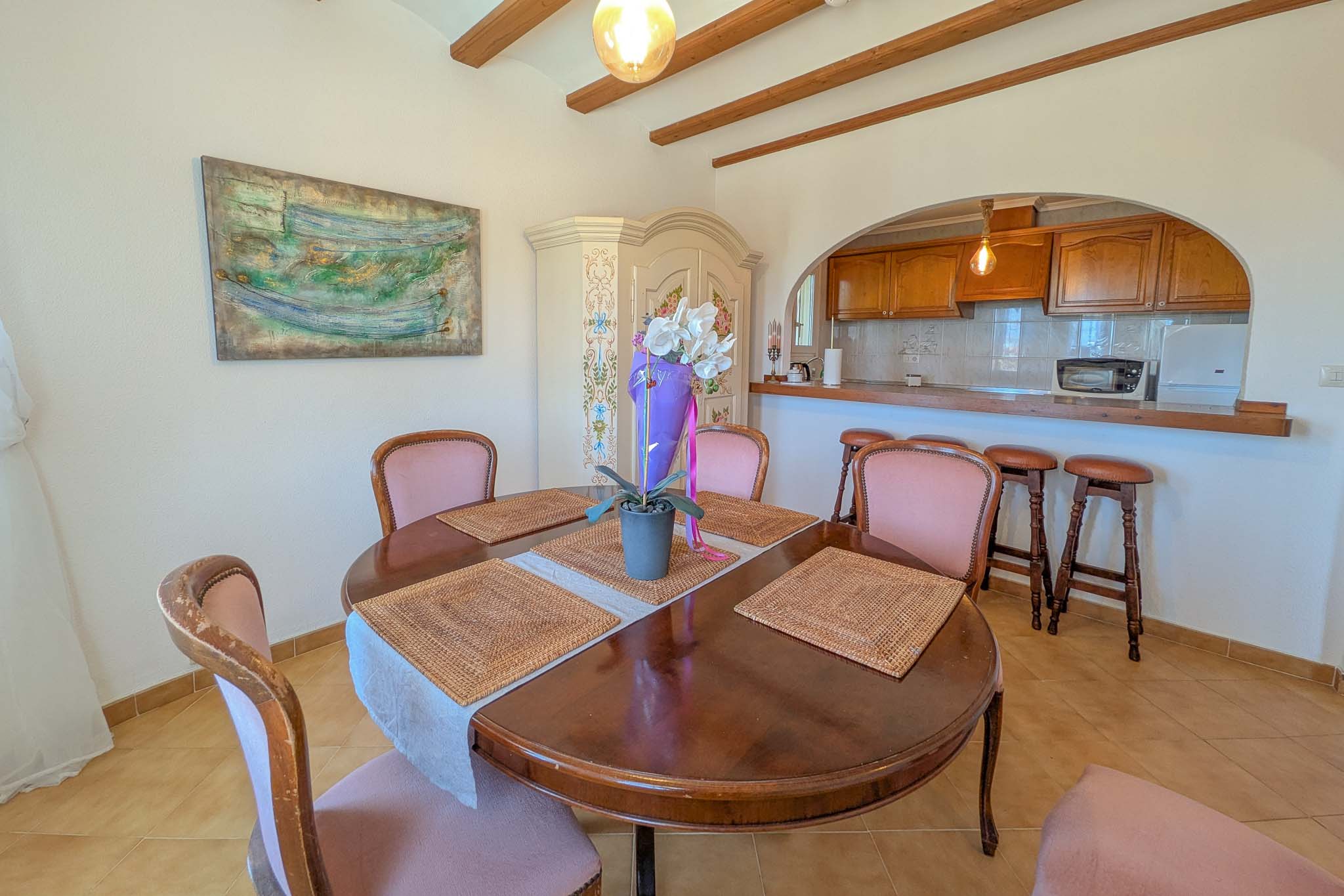 Dining area featuring a circular wooden table and exposed ceiling beams, integrated with a semi-open kitchen via an arched breakfast bar with stools.