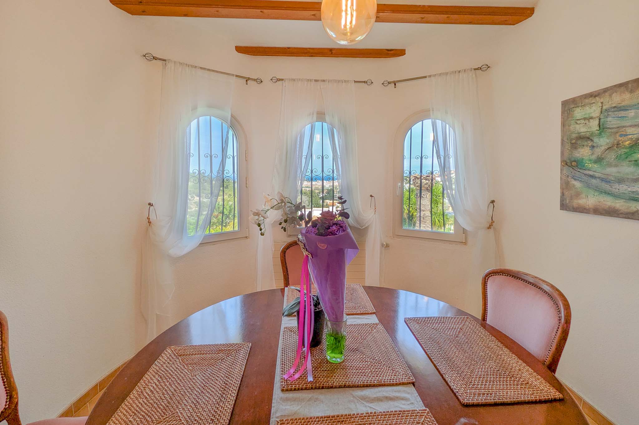 Dining area featuring a circular wooden table and three arched windows overlooking the sea. The room includes exposed wooden ceiling beams and natural lighting.