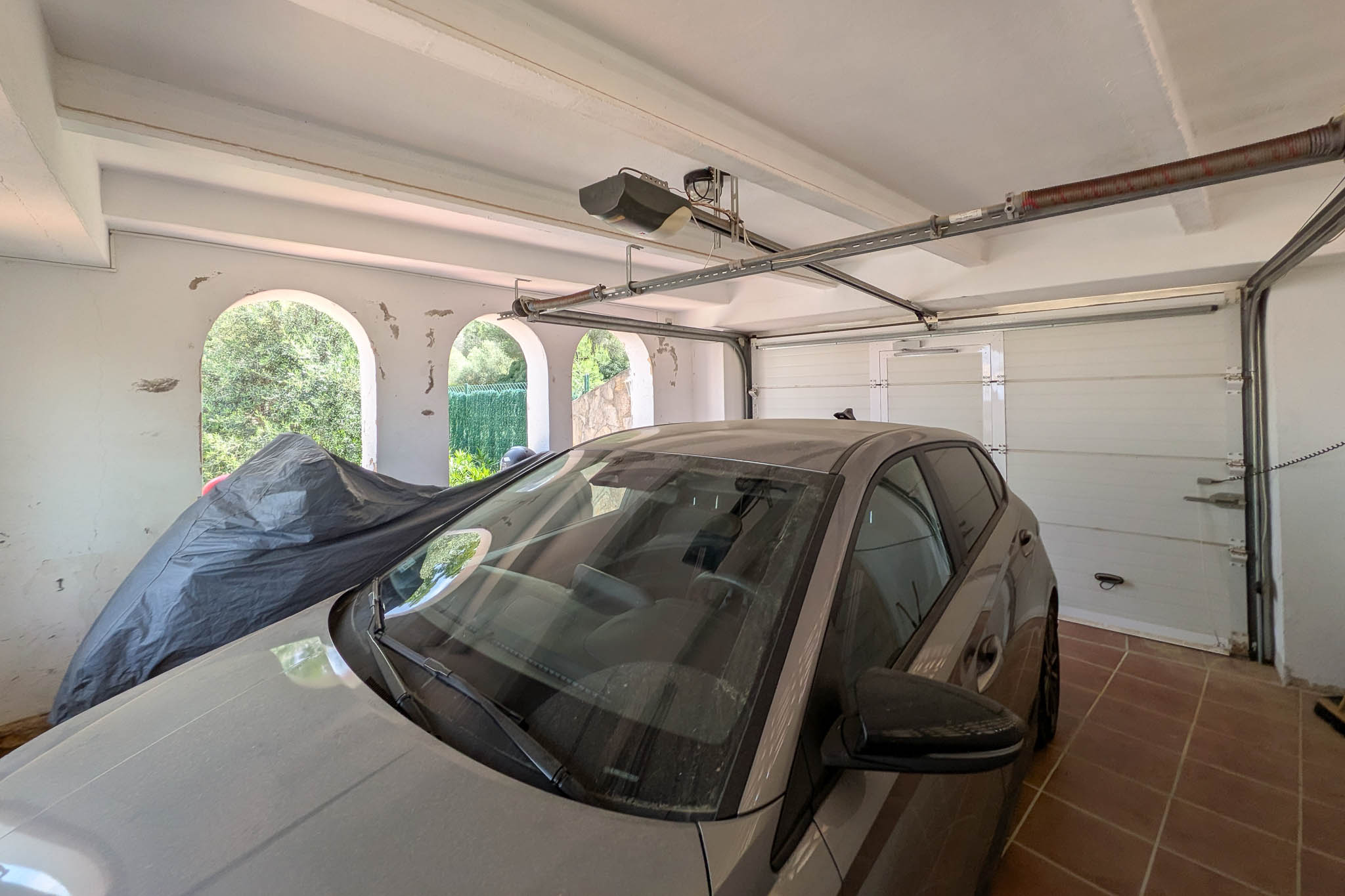 Enclosed garage equipped with an automatic sectional door, tiled flooring, and beamed ceilings. Arched side openings offer natural ventilation and light.