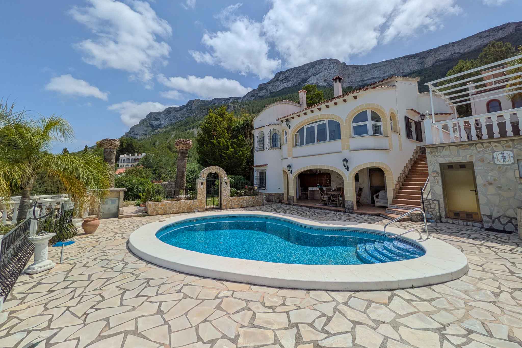 Exterior view featuring a kidney-shaped pool and stone-paved terrace, set against the white facade with arched details and the Montgó mountain backdrop.
