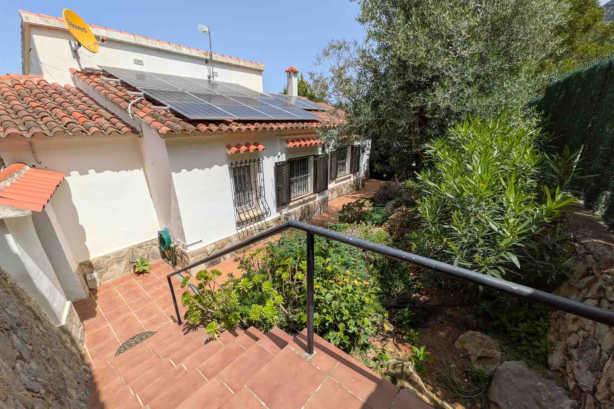 Exterior view showing the white facade with rooftop solar panels, terracotta stairs with metal railings, and integrated Mediterranean landscaping.