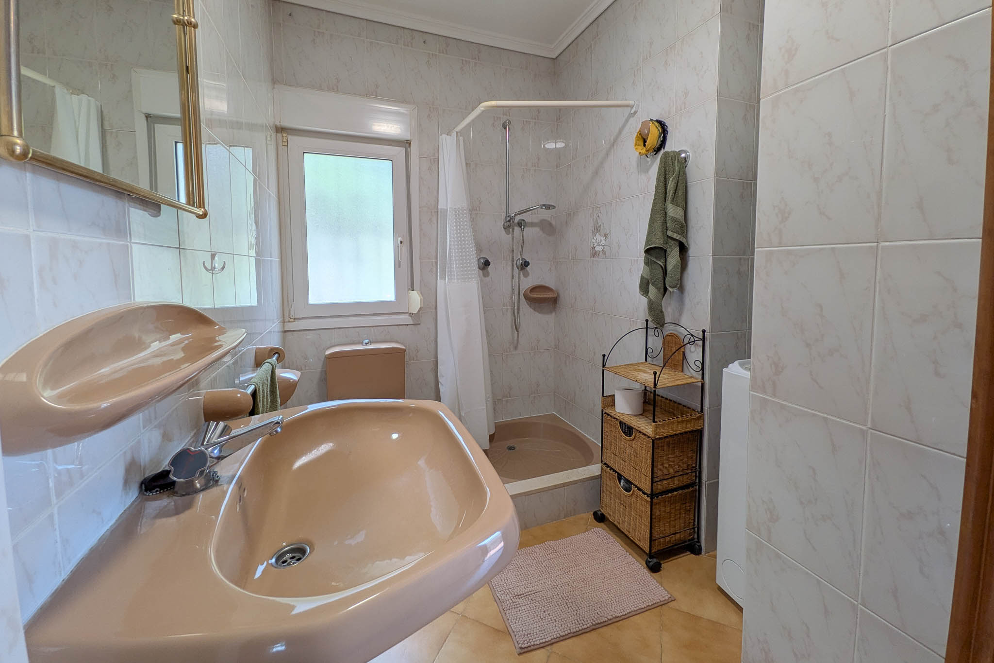 Functional bathroom featuring salmon-toned ceramics, a shower tray with curtain, tiled walls, and a double-glazed window for natural ventilation.