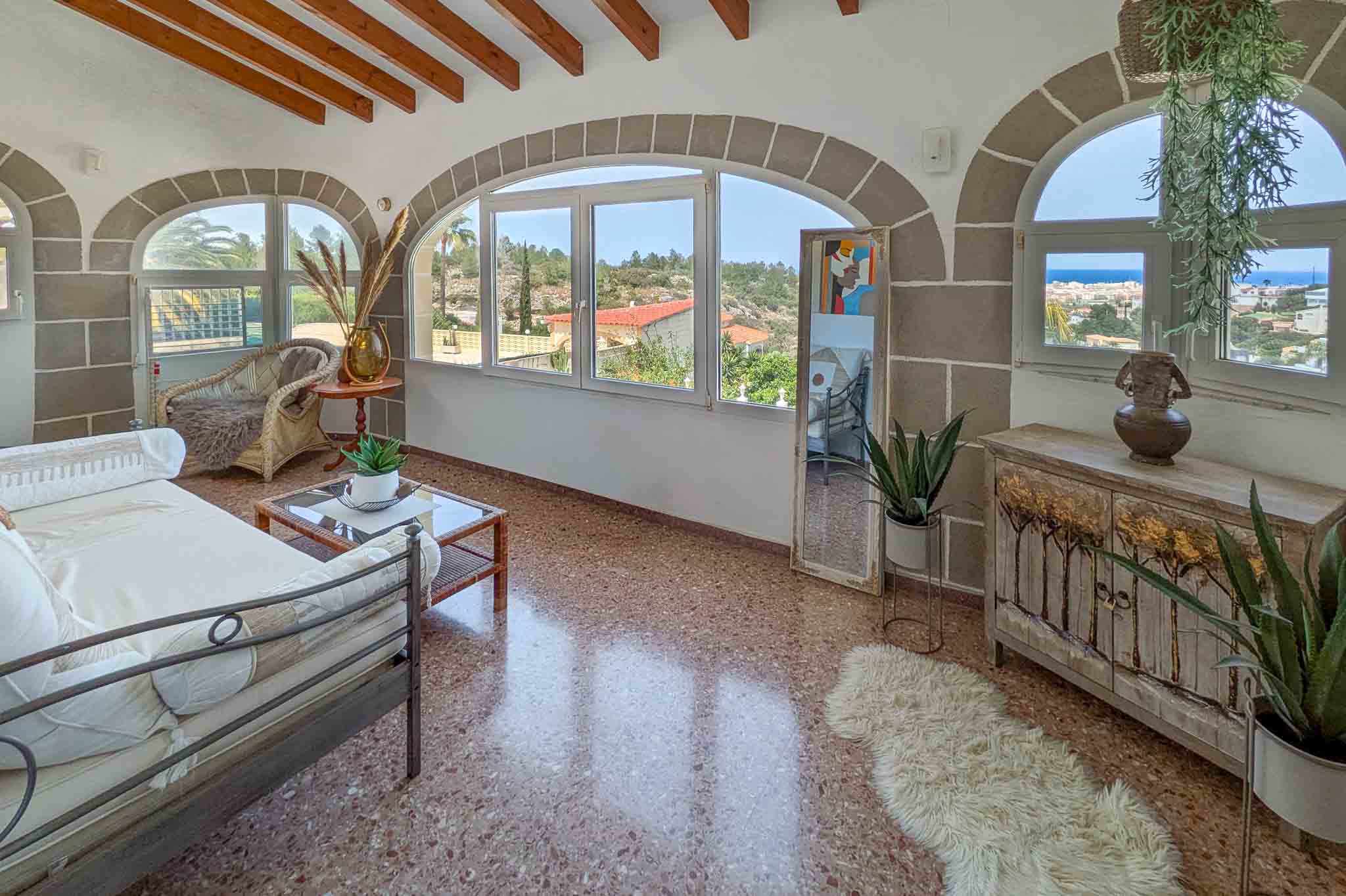 Glazed terrace featuring stone arches, exposed wooden beams, and terrazzo flooring. Large windows provide panoramic views of the Mediterranean Sea and Dénia.