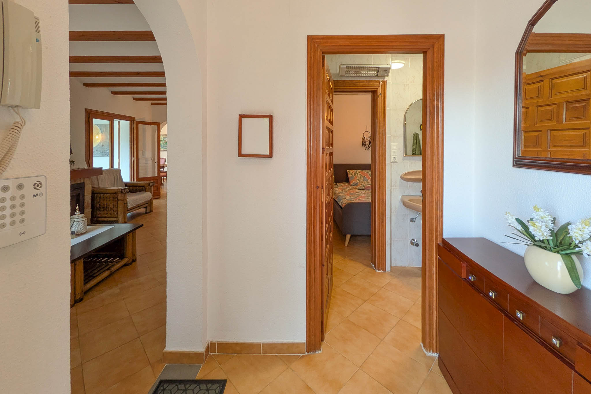 Hallway with terracotta tiling connecting the beamed living area to a bedroom and bathroom, featuring natural wood frames and traditional Mediterranean arches.