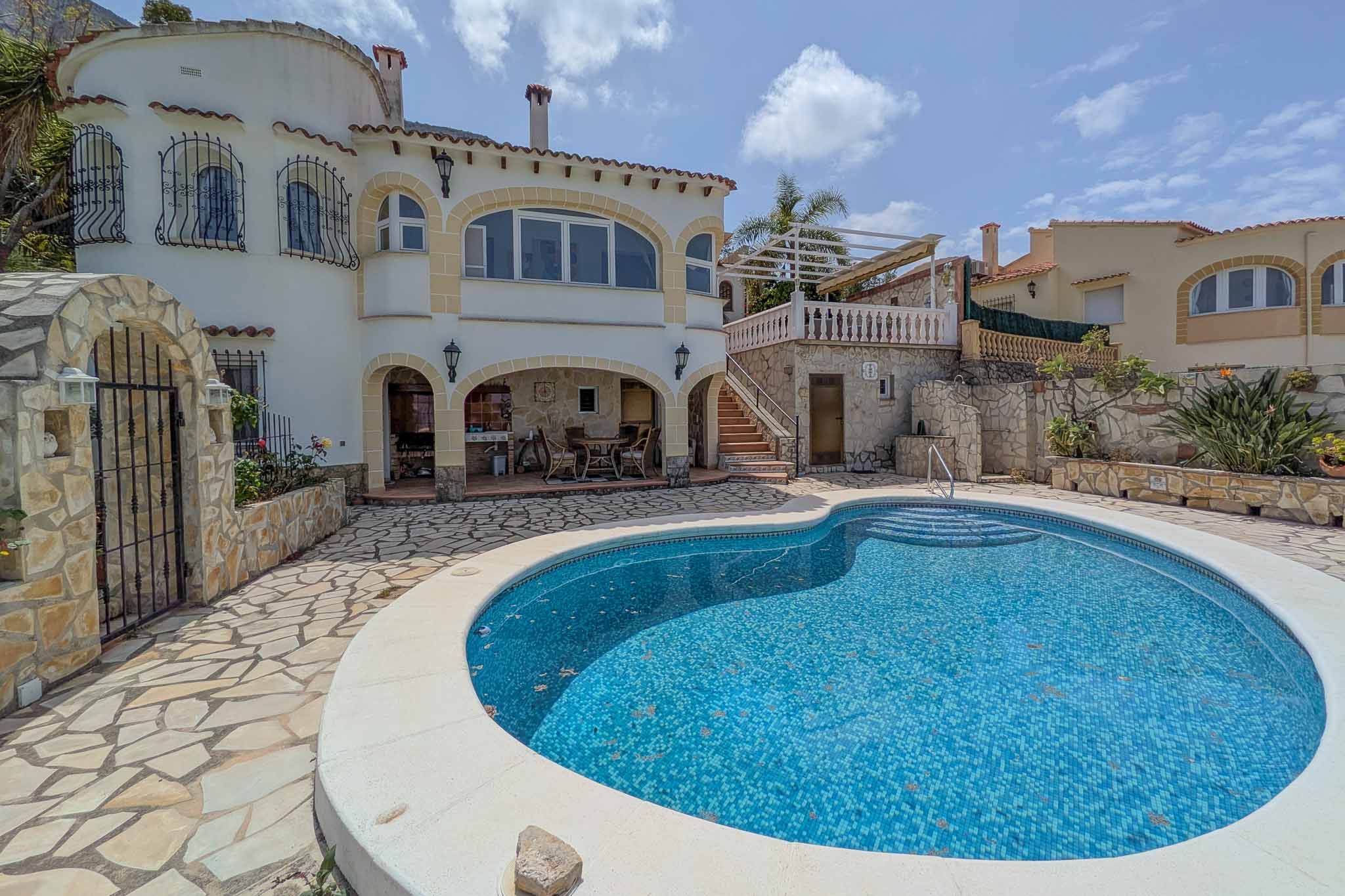 Kidney-shaped tiled pool surrounded by a natural stone terrace, featuring a covered porch with a summer kitchen and outdoor dining space.