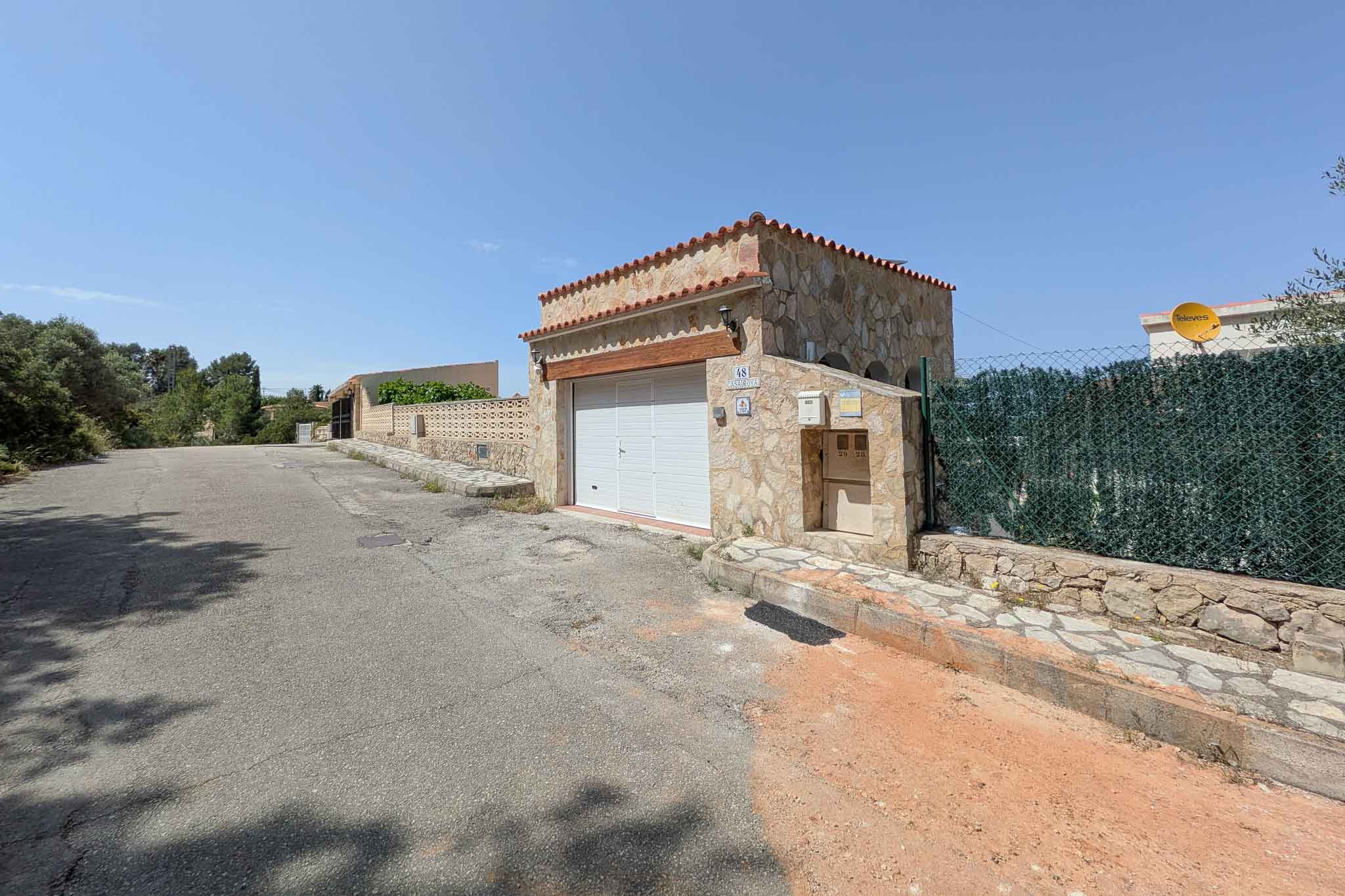 Main entrance featuring a stone-clad garage with a wooden lintel and terracotta roof, situated along a quiet residential street in La Pedrera.