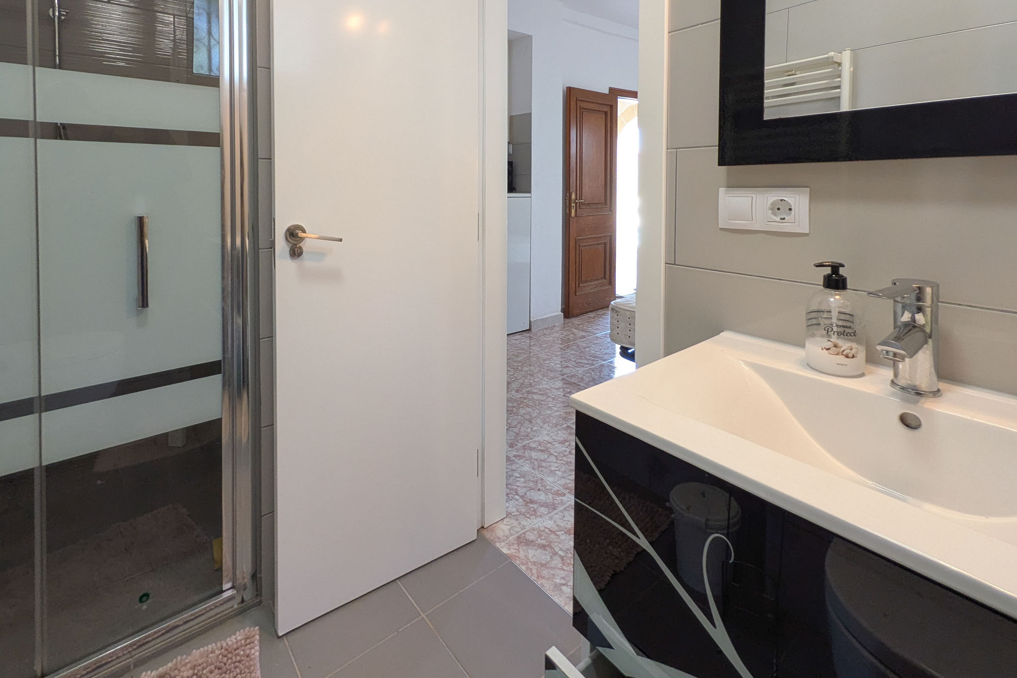 Modern bathroom featuring a black patterned vanity, glass shower enclosure, and grey tiling. The view extends through a white door to a hallway with classic tile flooring.