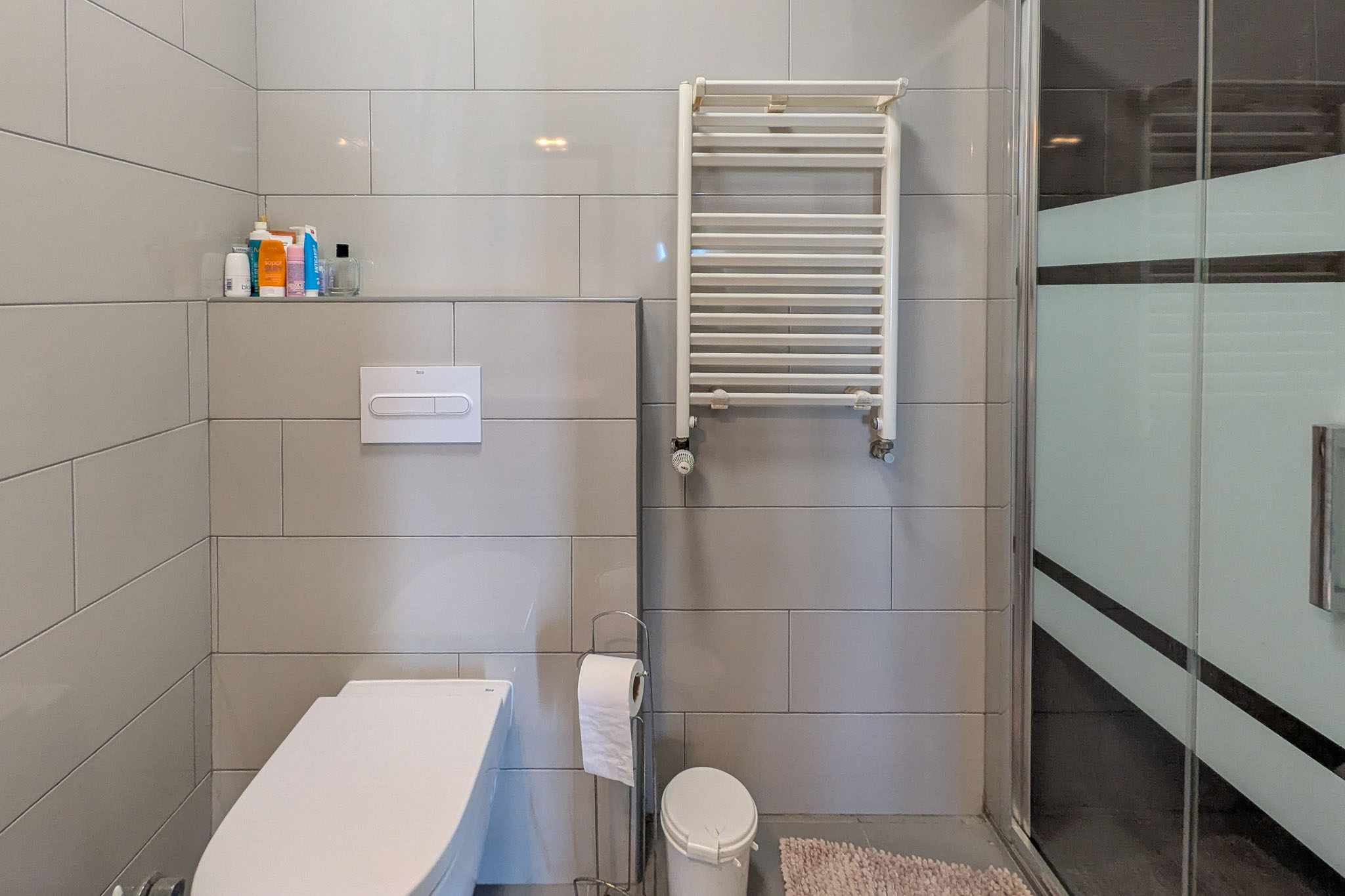 Modern bathroom featuring a wall-hung toilet, grey ceramic tiling, a white heated towel rail, and a glass shower enclosure with a frosted privacy band.