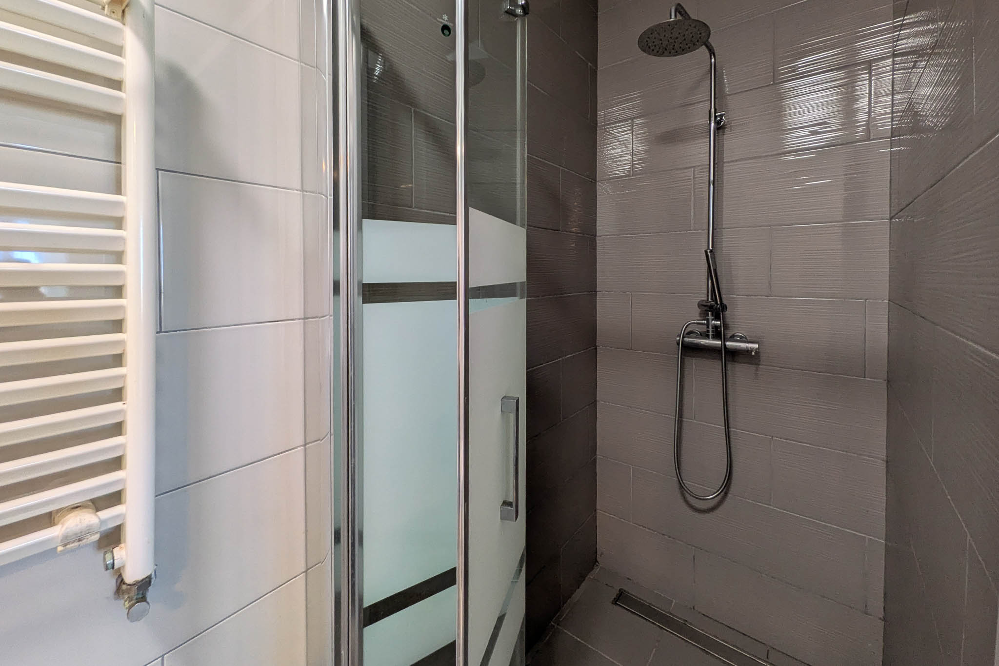 Modern walk-in shower with textured grey wall tiles, glass enclosure with frosted privacy bands, chrome rainfall showerhead, and a white heated towel rail.