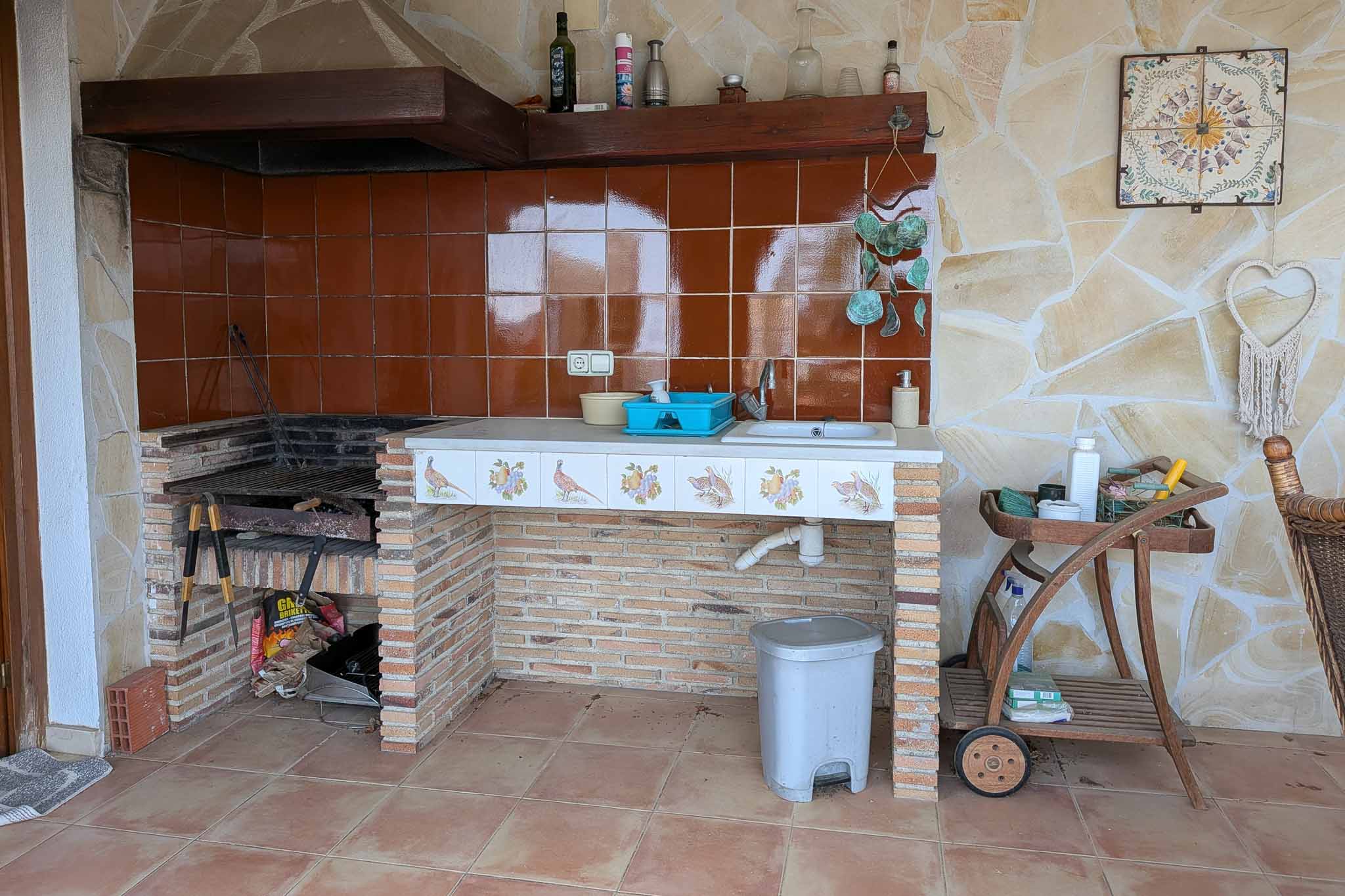 Outdoor summer kitchen featuring a built-in brick barbecue, integrated sink, and decorative tiled countertop against a natural stone masonry wall.