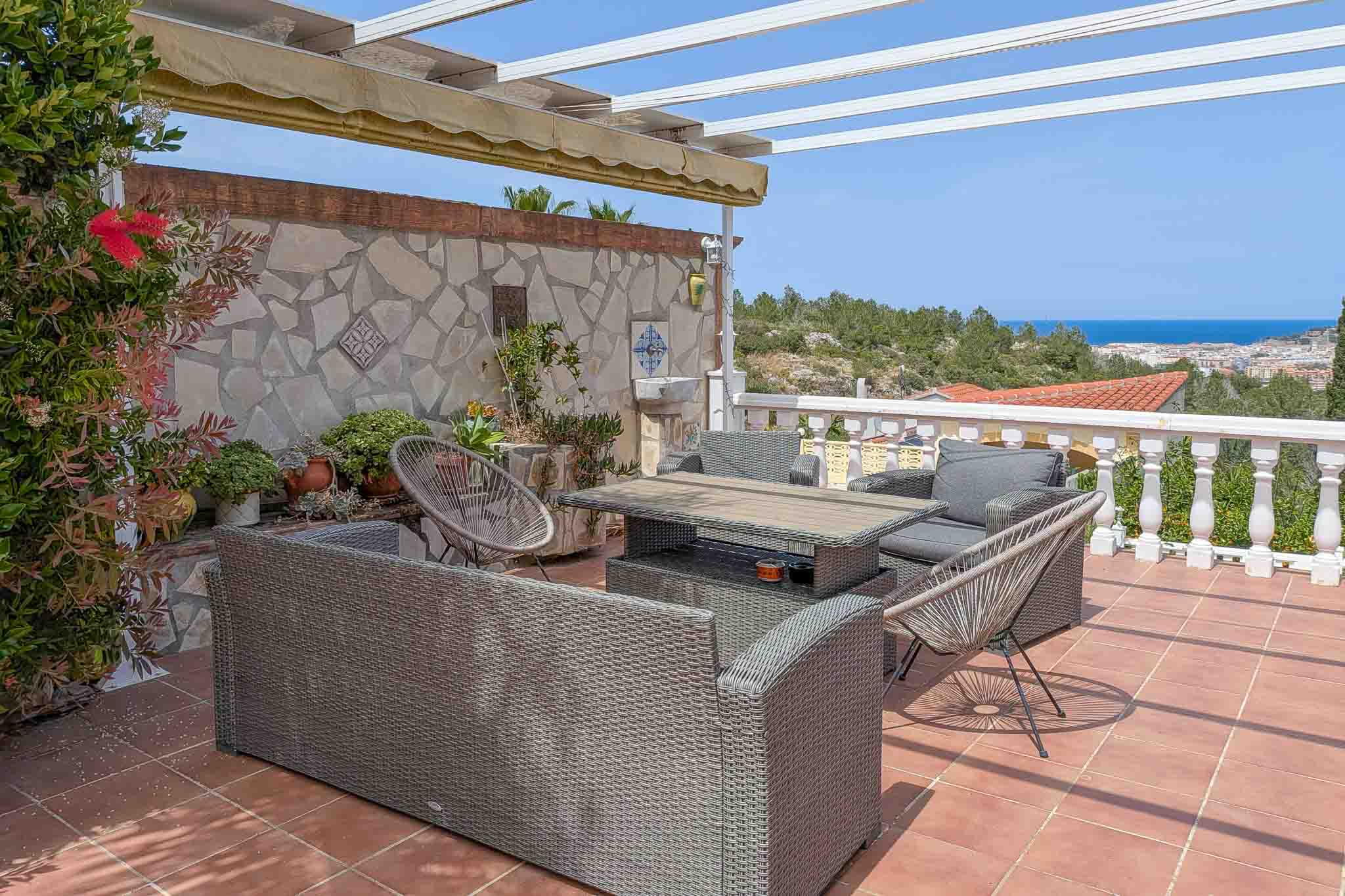 Outdoor terrace featuring rattan seating, terracotta flooring, and panoramic sea views. Includes a pergola with retractable awning and a decorative natural stone wall.