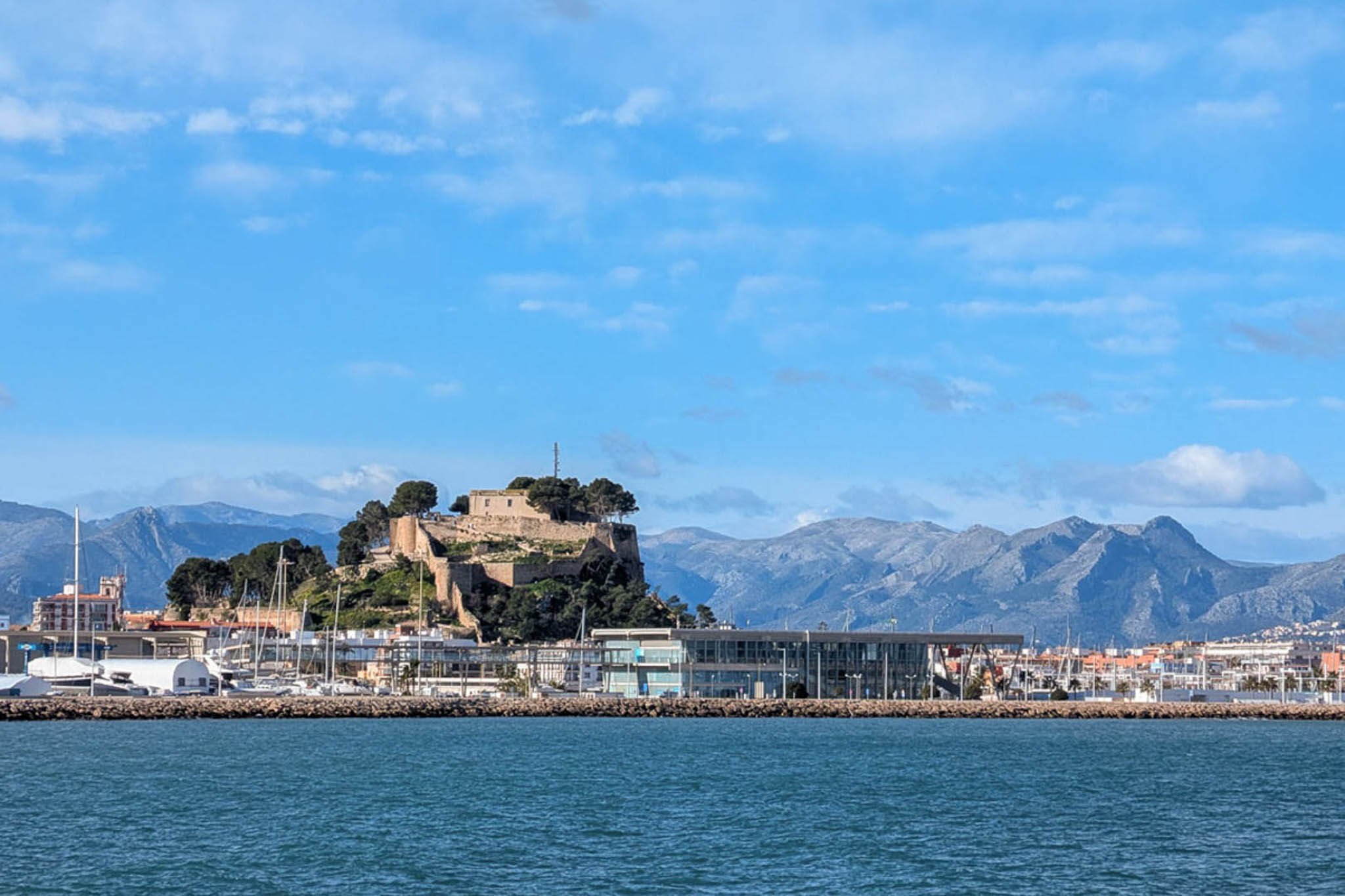 Panoramic view of Dénia Castle and the marina from the sea, showing the historic fortress, the modern port terminal, and the mountain skyline.
