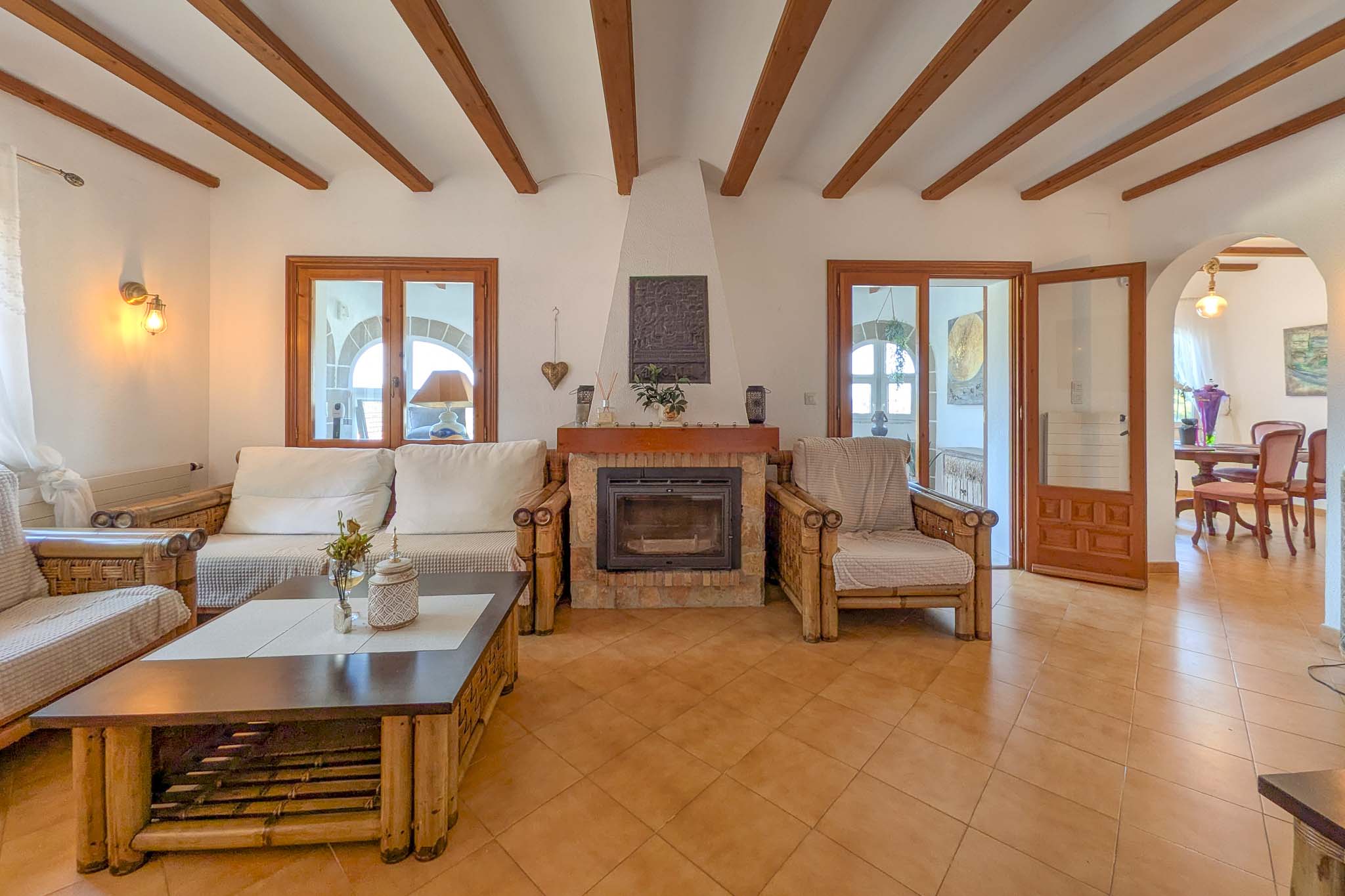 Rustic living room featuring a stone fireplace, exposed wooden beams, and terracotta flooring. Bamboo furniture and arched doorways connect the space to the dining area.