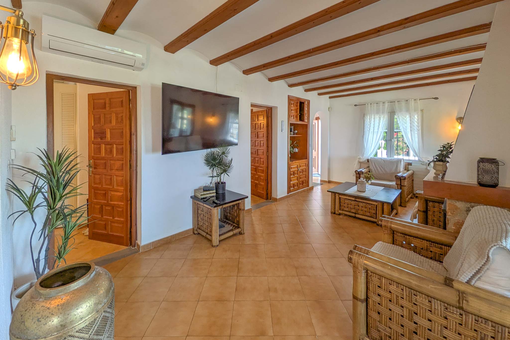 Rustic living room featuring exposed ceiling beams, terracotta flooring, and bamboo furniture. Equipped with split-system air conditioning and traditional carved wooden doors.