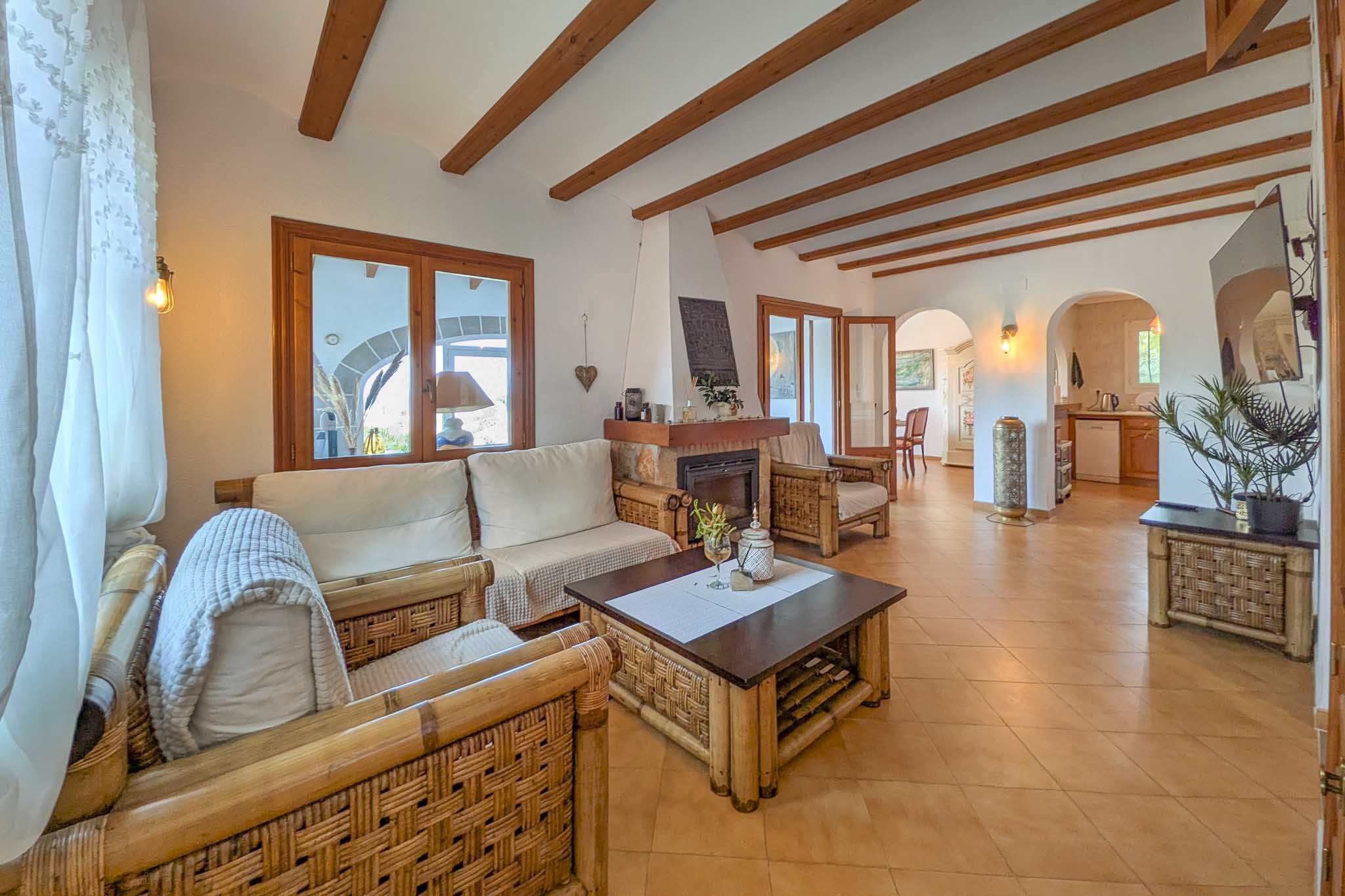 Rustic living room featuring exposed wooden ceiling beams, a stone fireplace, and bamboo furniture. Arched doorways lead to the dining area and kitchen.
