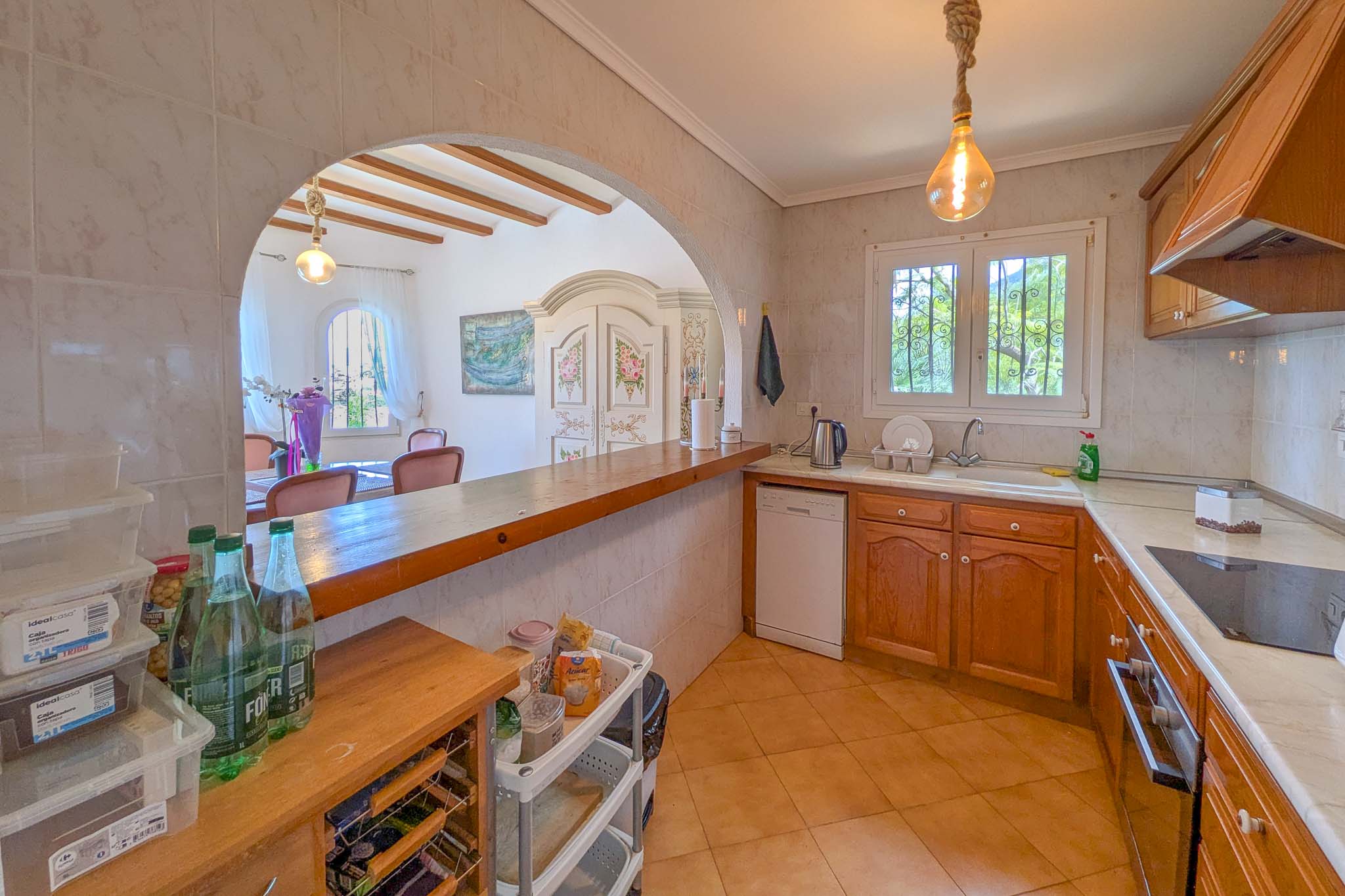 Semi-open kitchen featuring wooden cabinetry, a solid wood breakfast bar, and an arched opening to the dining area with exposed ceiling beams.