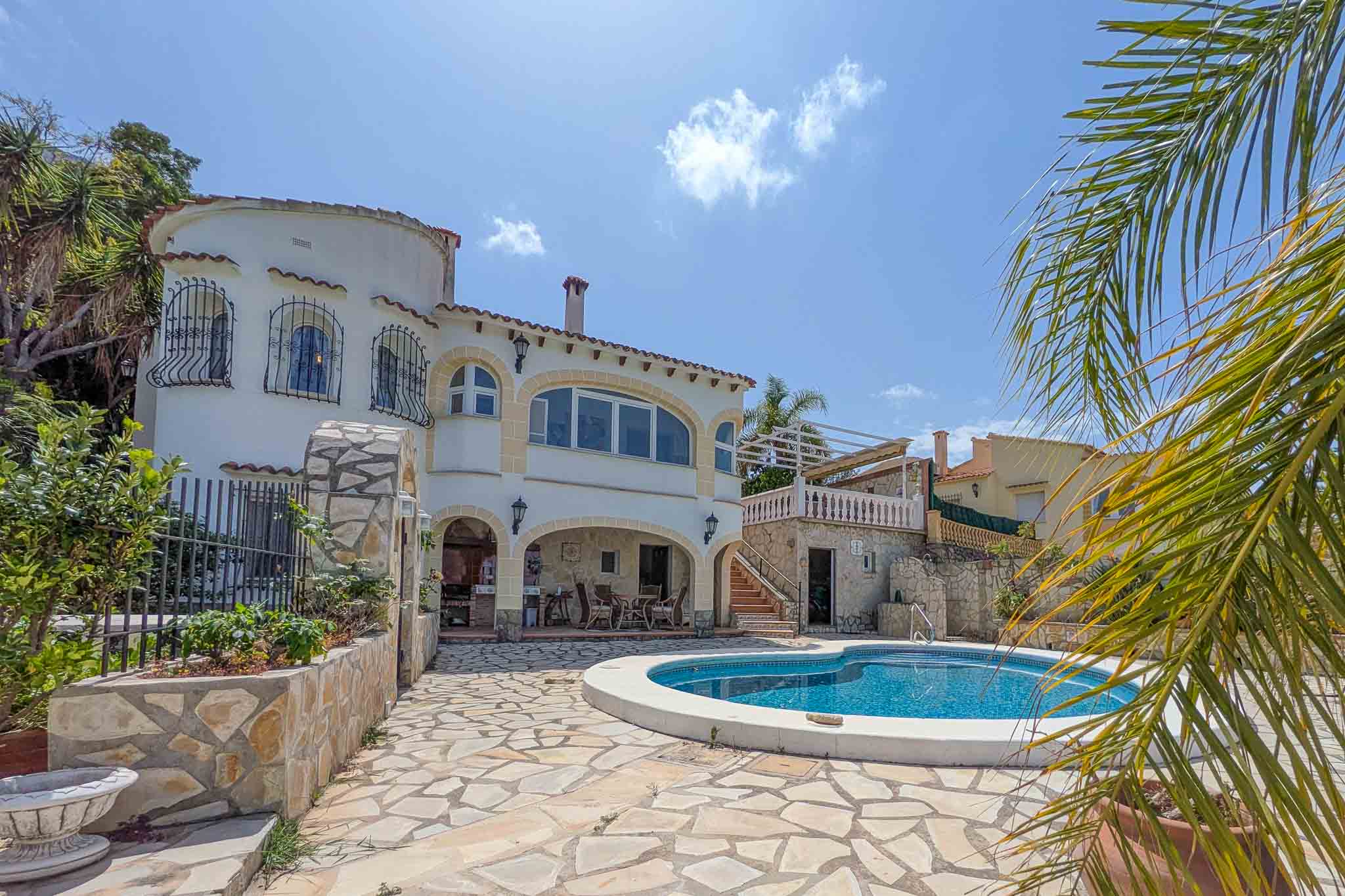 Villa exterior featuring a curved swimming pool, natural stone terrace, and a covered porch with a summer kitchen. The white facade displays arched windows and a turret.