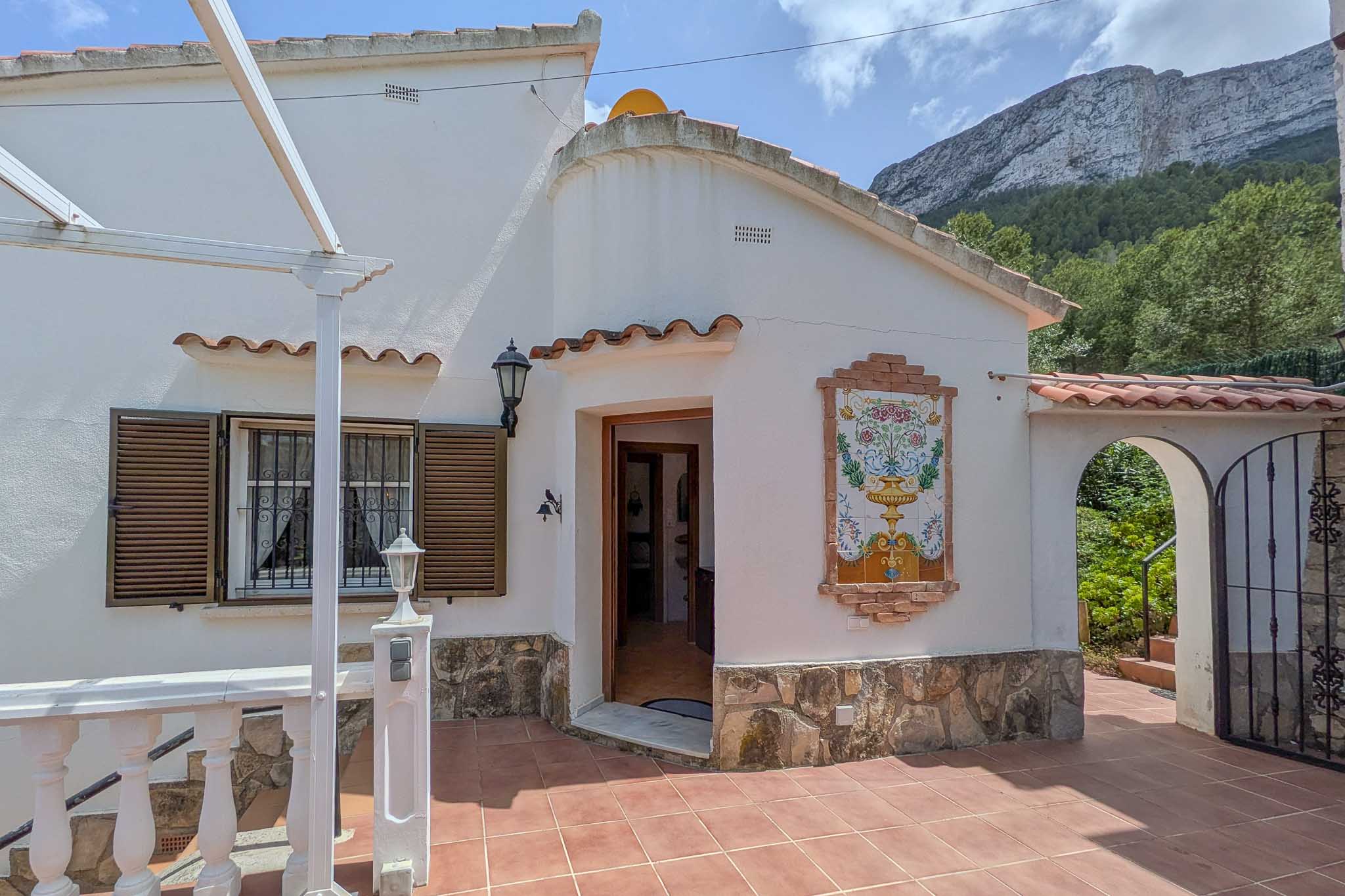 White facade featuring a decorative ceramic mural, stone base, and terracotta tiling. The Montgó mountain provides a natural backdrop to the entrance.