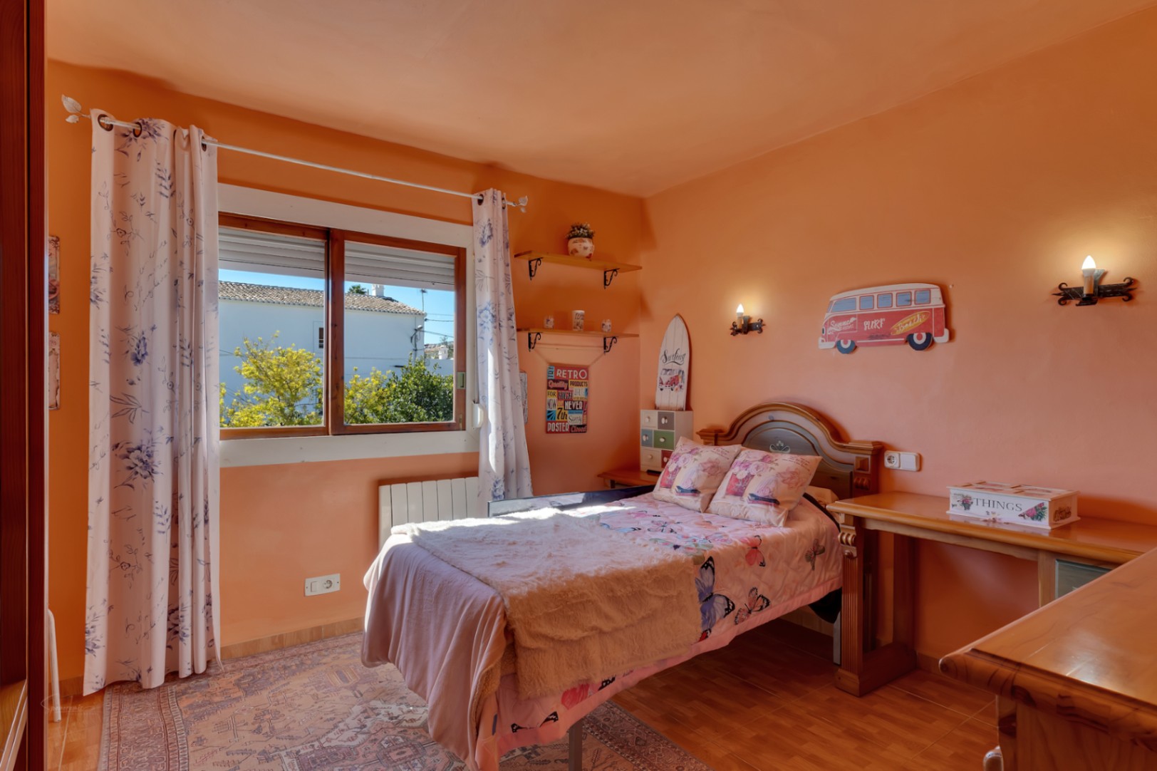 Bright bedroom featuring peach-colored walls, wooden furniture, an integrated desk, and a window. It includes a wall radiator and traditional tiled flooring.