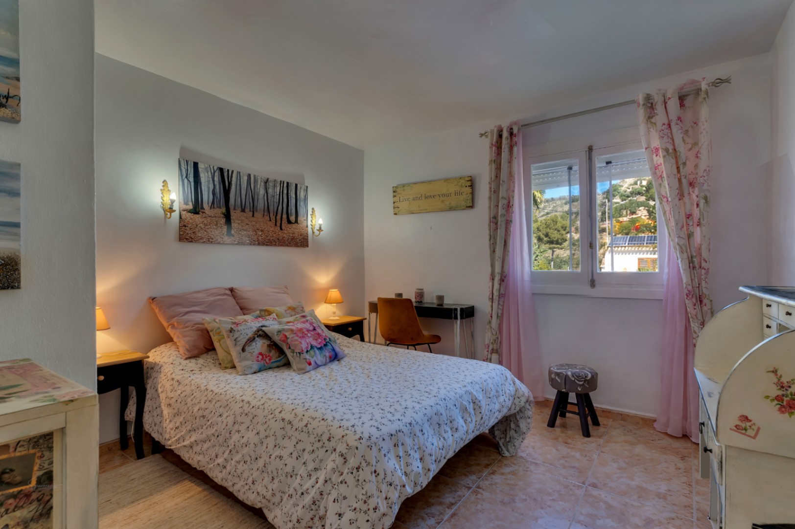 Bright bedroom featuring tiled flooring, a functional workspace, and a window offering views of the surrounding hills. Decorated with classic furniture and warm lighting.